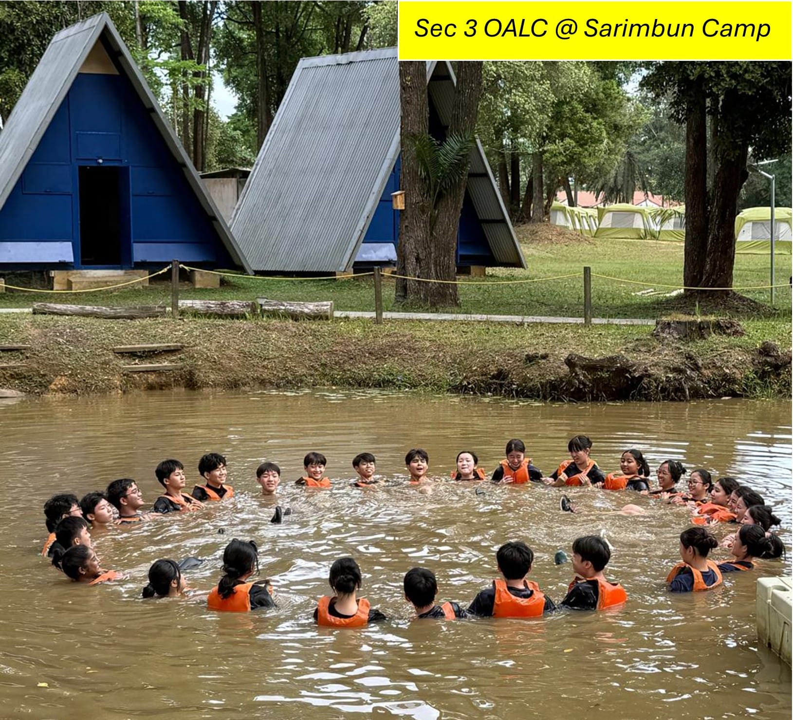 Students in orange life vests form a circle in a muddy pond at Sarimbun Camp, with blue A-frame cabins behind them.