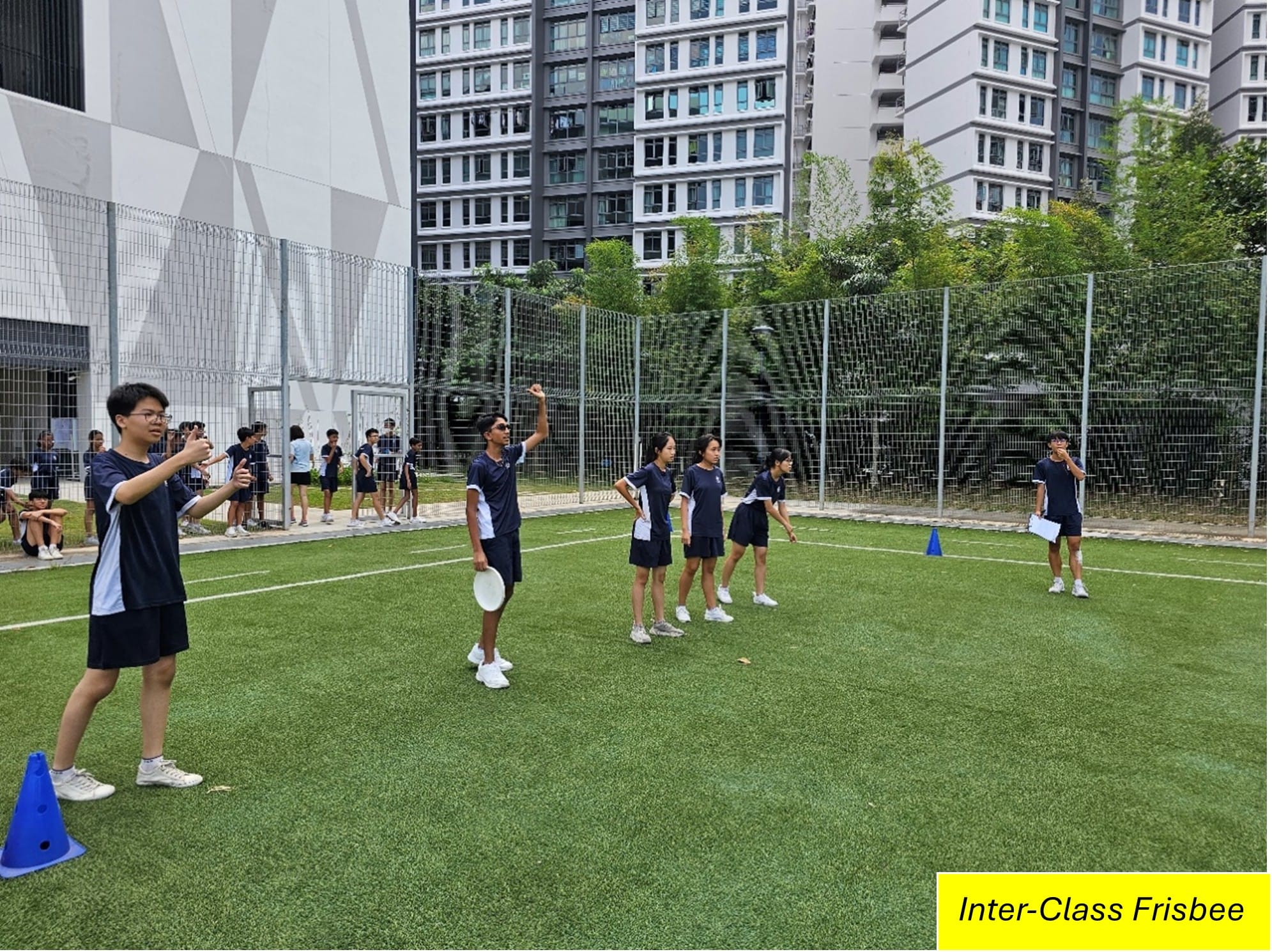 Students in navy uniforms play frisbee on an outdoor artificial turf field surrounded by tall buildings.
