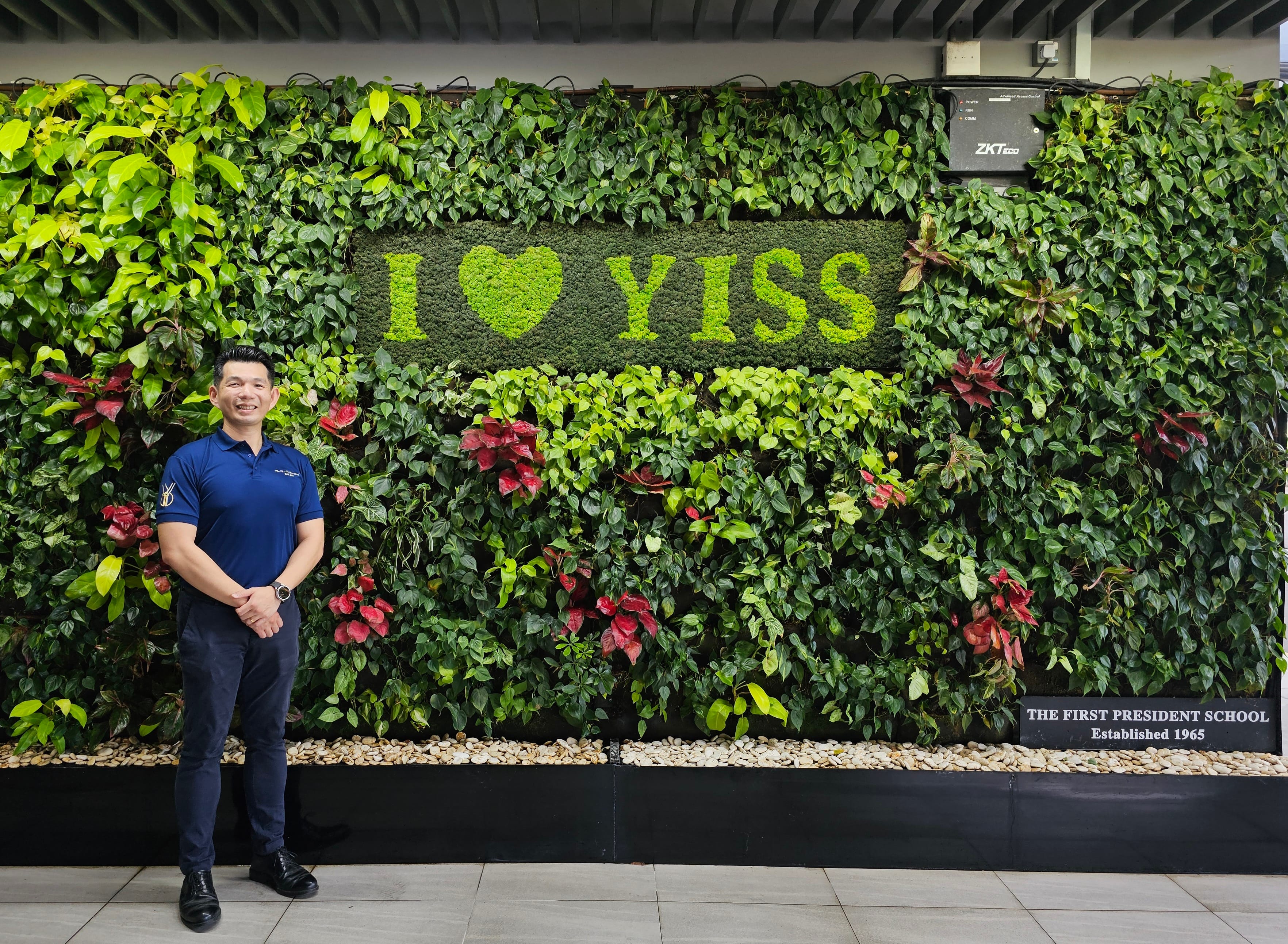 Man stands by a lush plant wall with I ♥ YISS and a plaque reading THE FIRST PRESIDENT SCHOOL Established 1965.