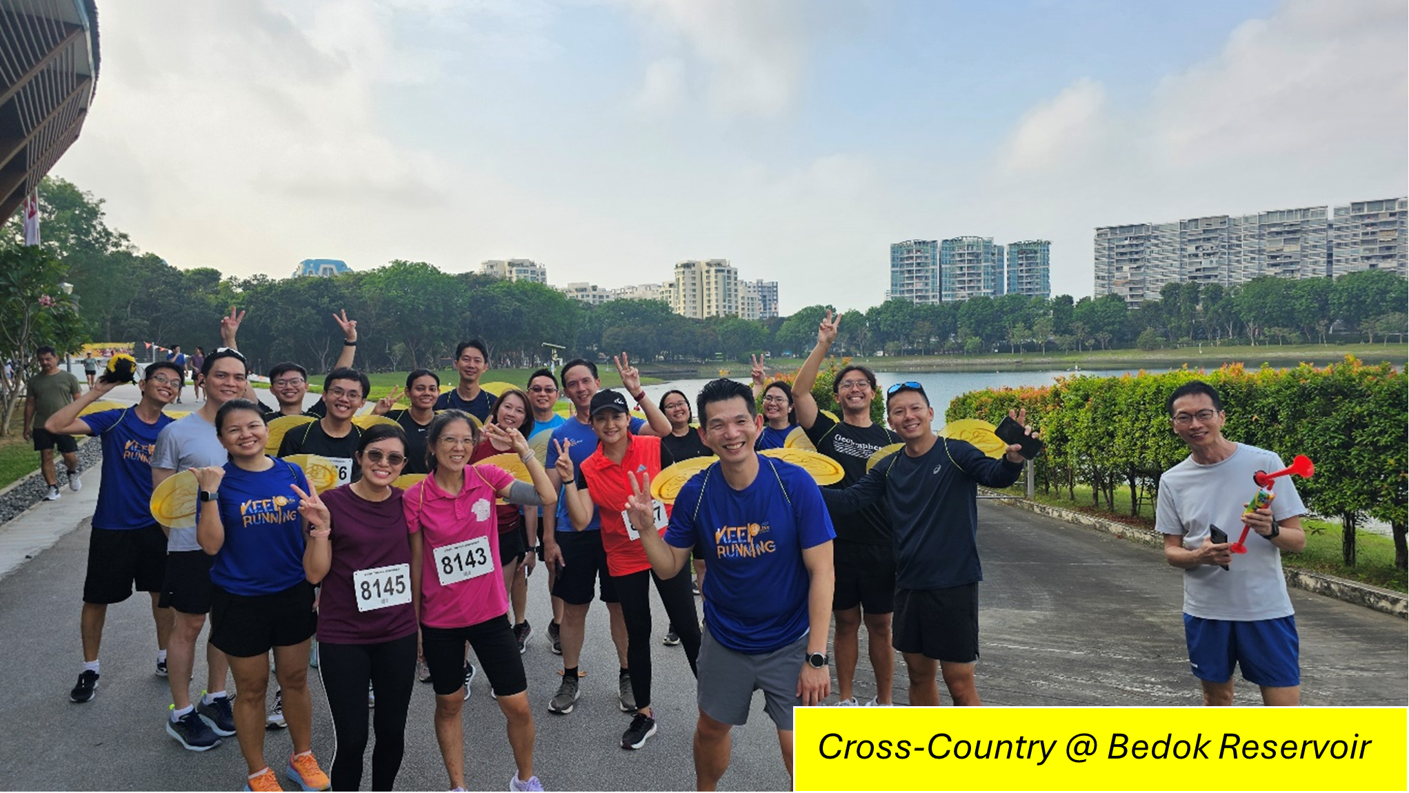 Group of runners posing with peace signs on a reservoir path, with city buildings in the background.