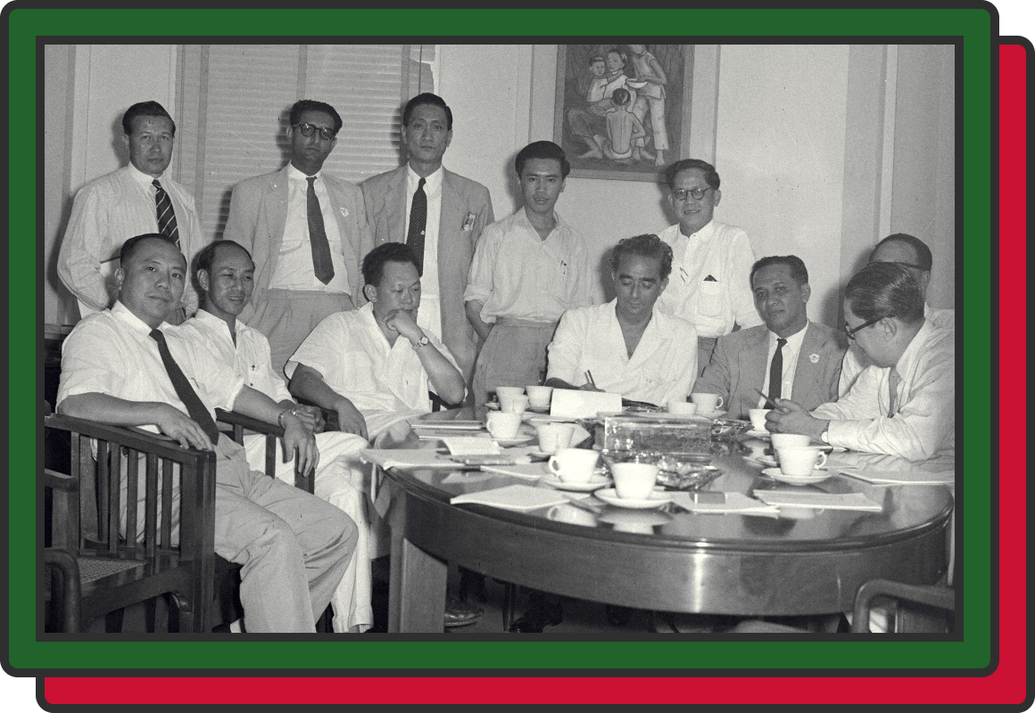 Black and white vintage photo of a group of men in formal attire seated and standing around a round table with teacups.