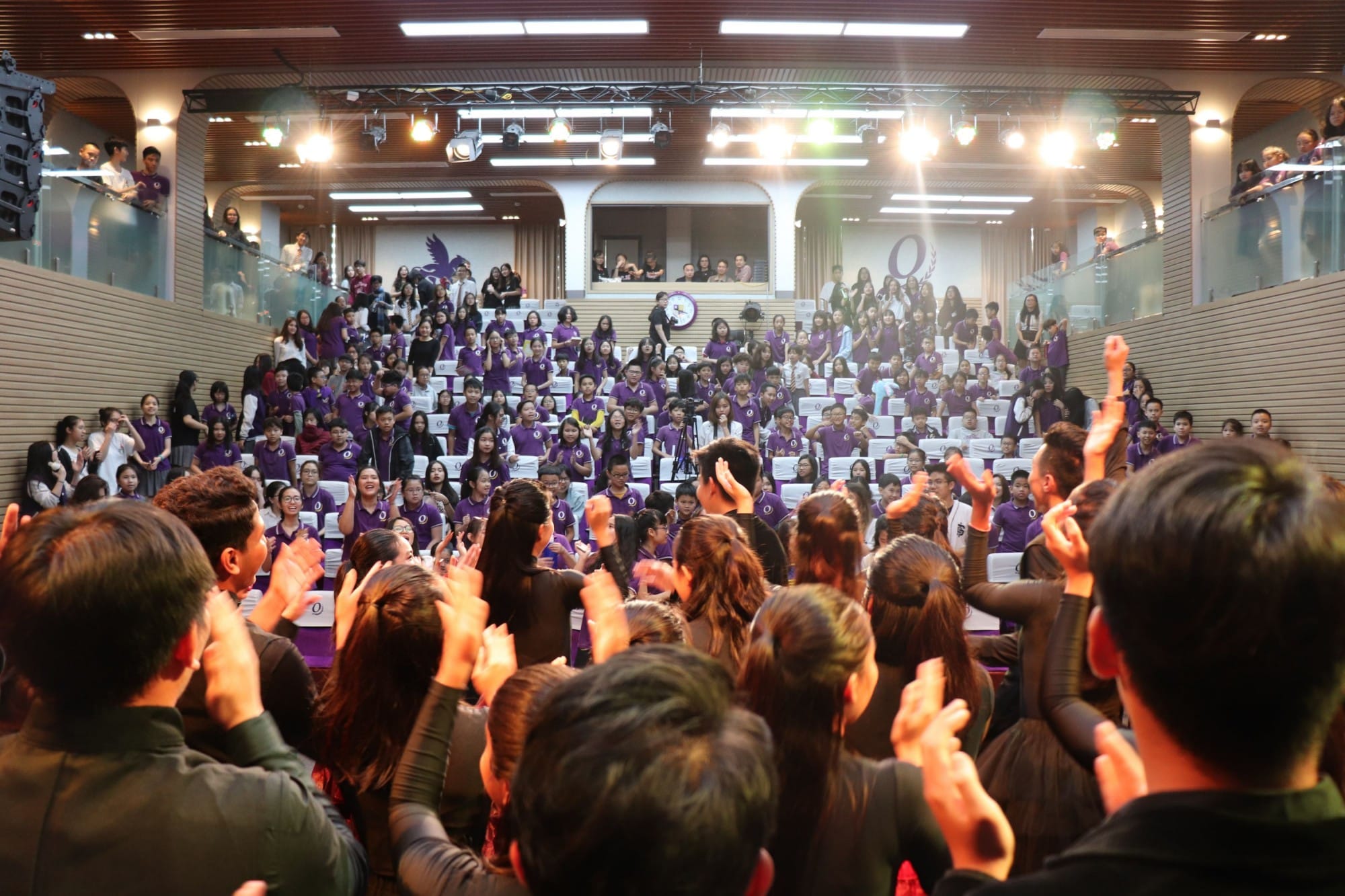 Large group of students in purple uniforms filling a lit auditorium, with hands raised and clapping.