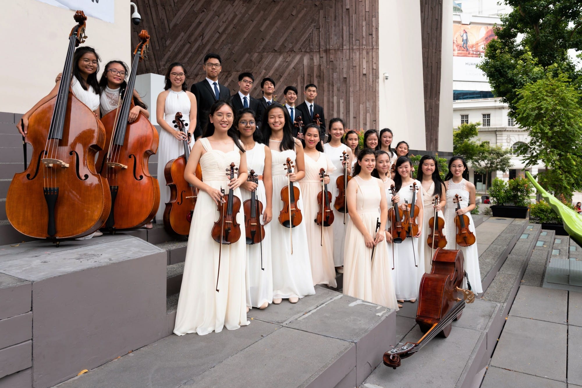 Young string orchestra members in white dresses and black suits pose outdoors holding violins and double basses.