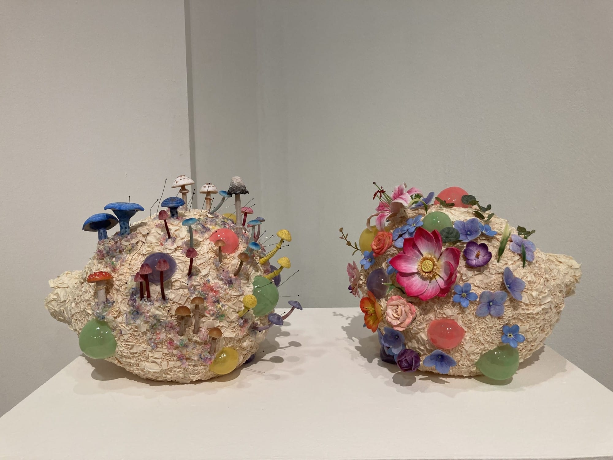 Two white textured sculptures shaped like hedgehogs, one covered in colorful mushrooms and one with bright flowers.