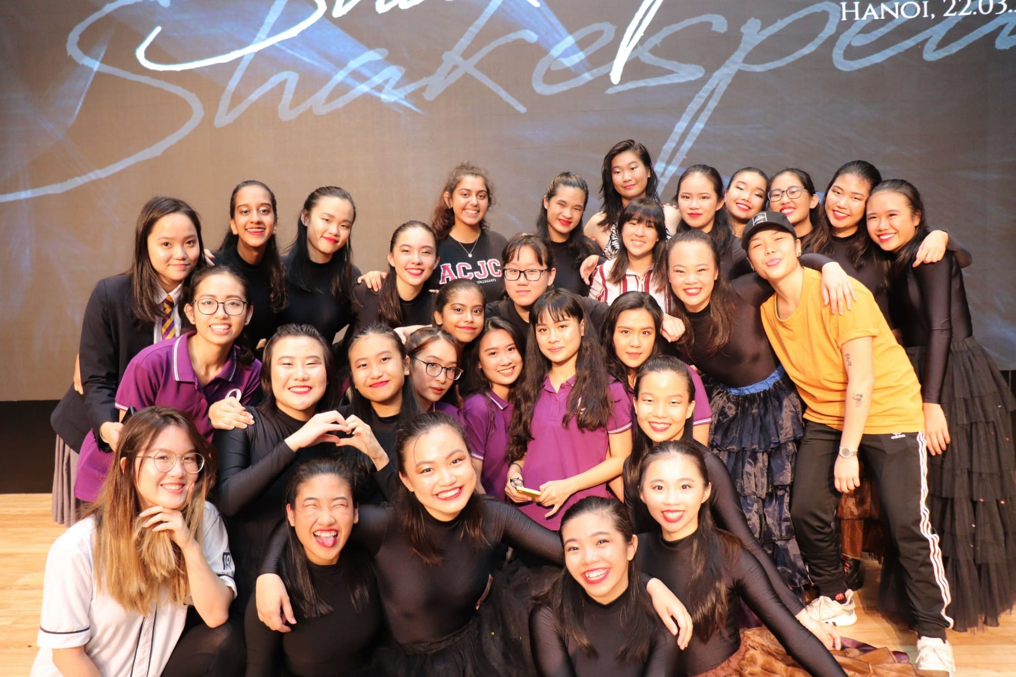 A large group of young women smiling on a stage in Hanoi, many wearing black outfits, with a Shakespeare backdrop.