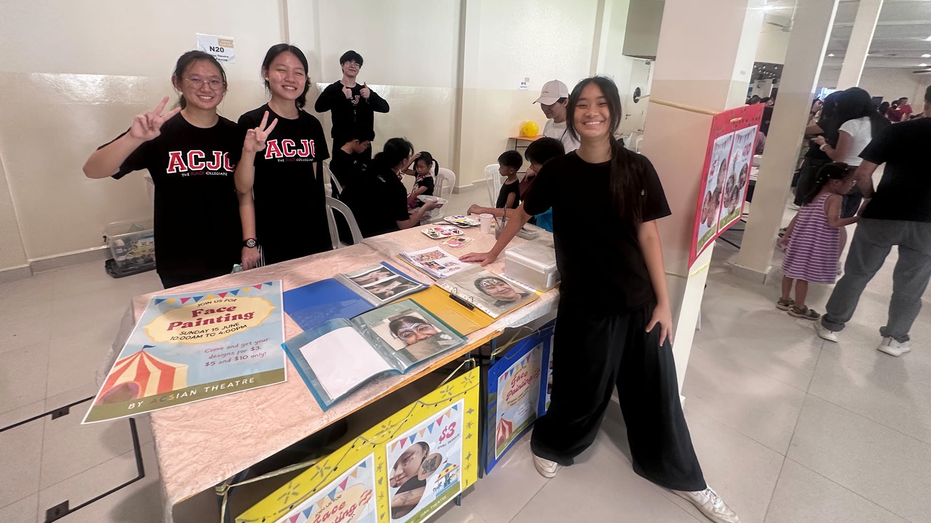 Three students in black ACJC shirts pose at a face painting booth with posters and sample photos at an event.