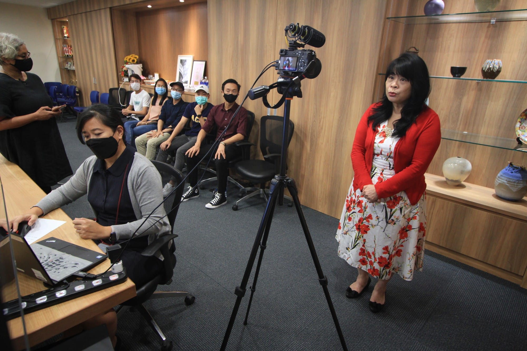 A woman in a red cardigan stands before a camera on a tripod while masked people sit and observe indoors.