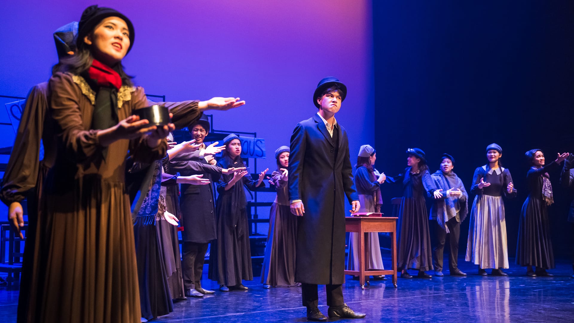 Stage performers in period costumes act on a blue-lit stage, with a woman in a red scarf and a man in a top hat.