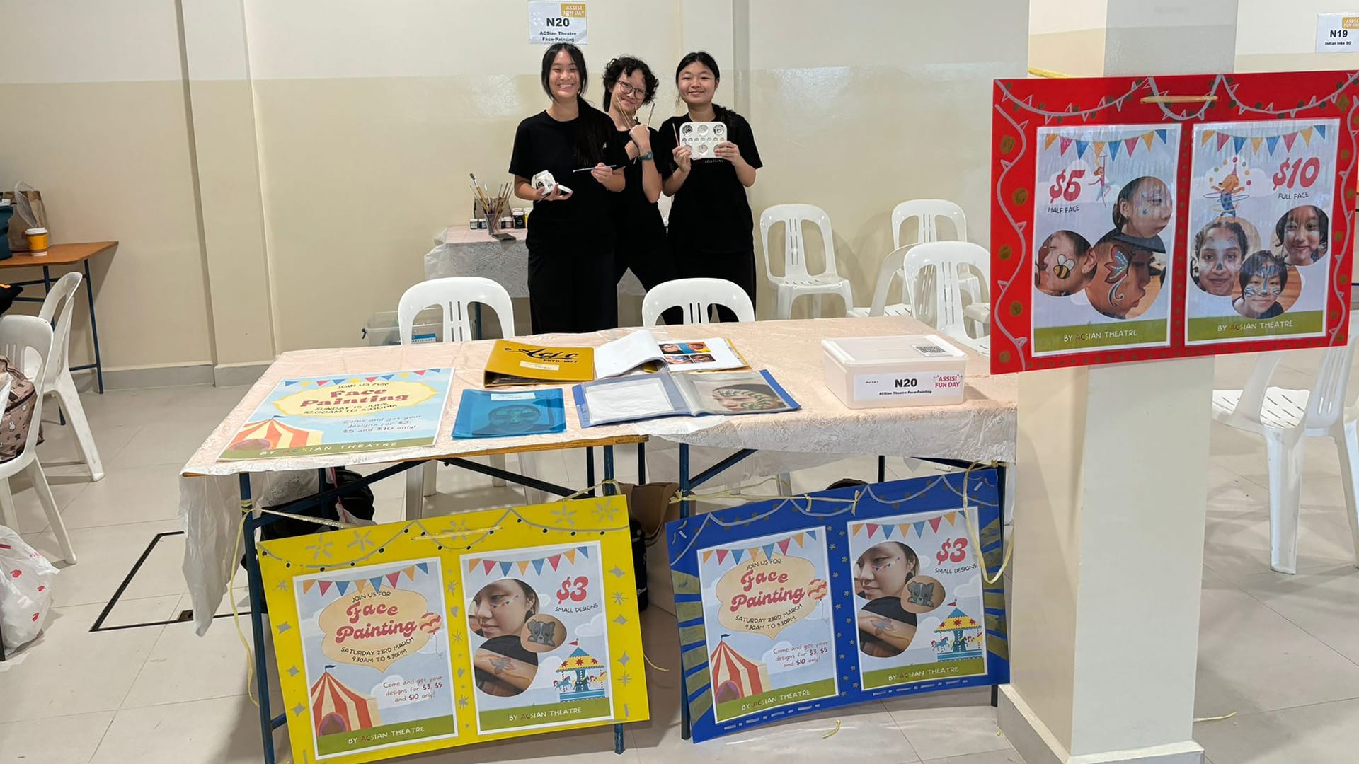 Three people in black stand behind a face painting booth with colorful posters showing prices of $3, $5, and $10.