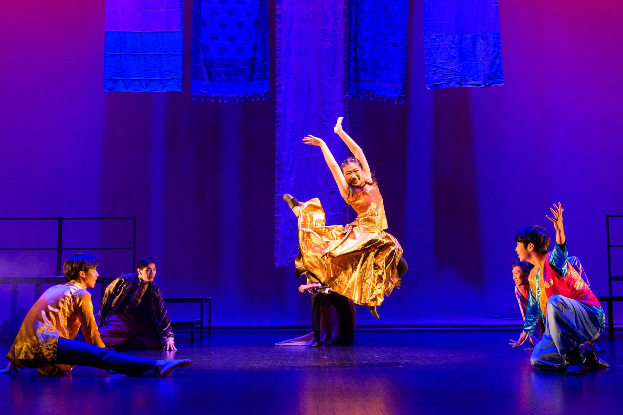 A woman in a gold dress leaps on a purple-lit stage while five performers in colorful costumes watch around her.