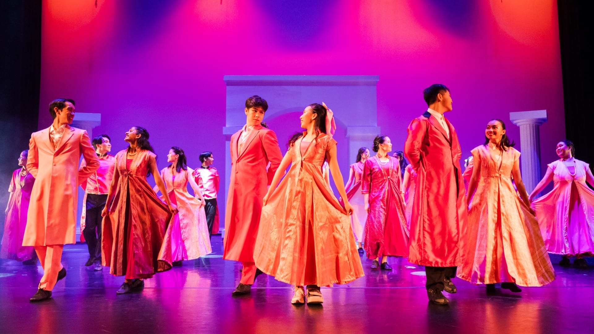 Performers in pink and red gowns and suits dance on a stage lit with pink and purple lighting.