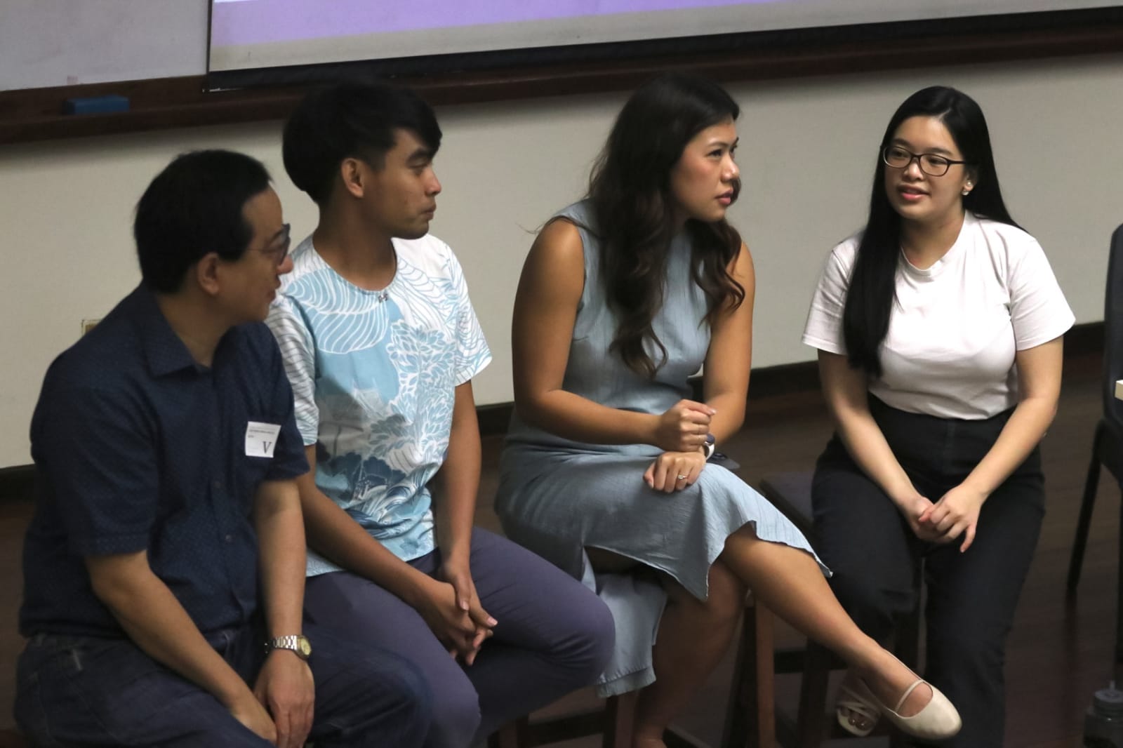 Four people sit in a row during a panel discussion, with a woman in a blue dress speaking to the group.