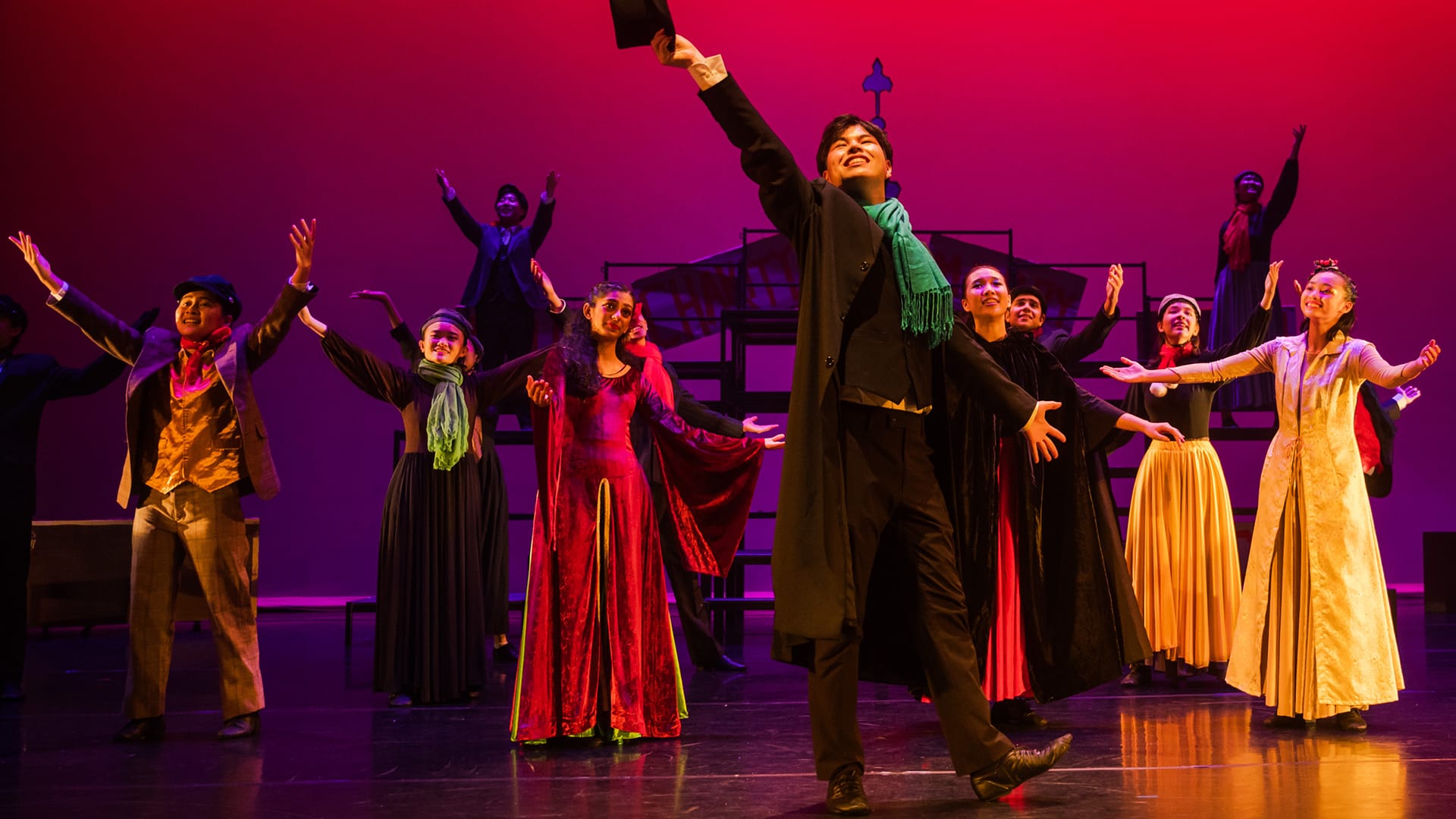 A group of performers in period costumes dance on a red-lit stage, with a lead male tossing a hat overhead.