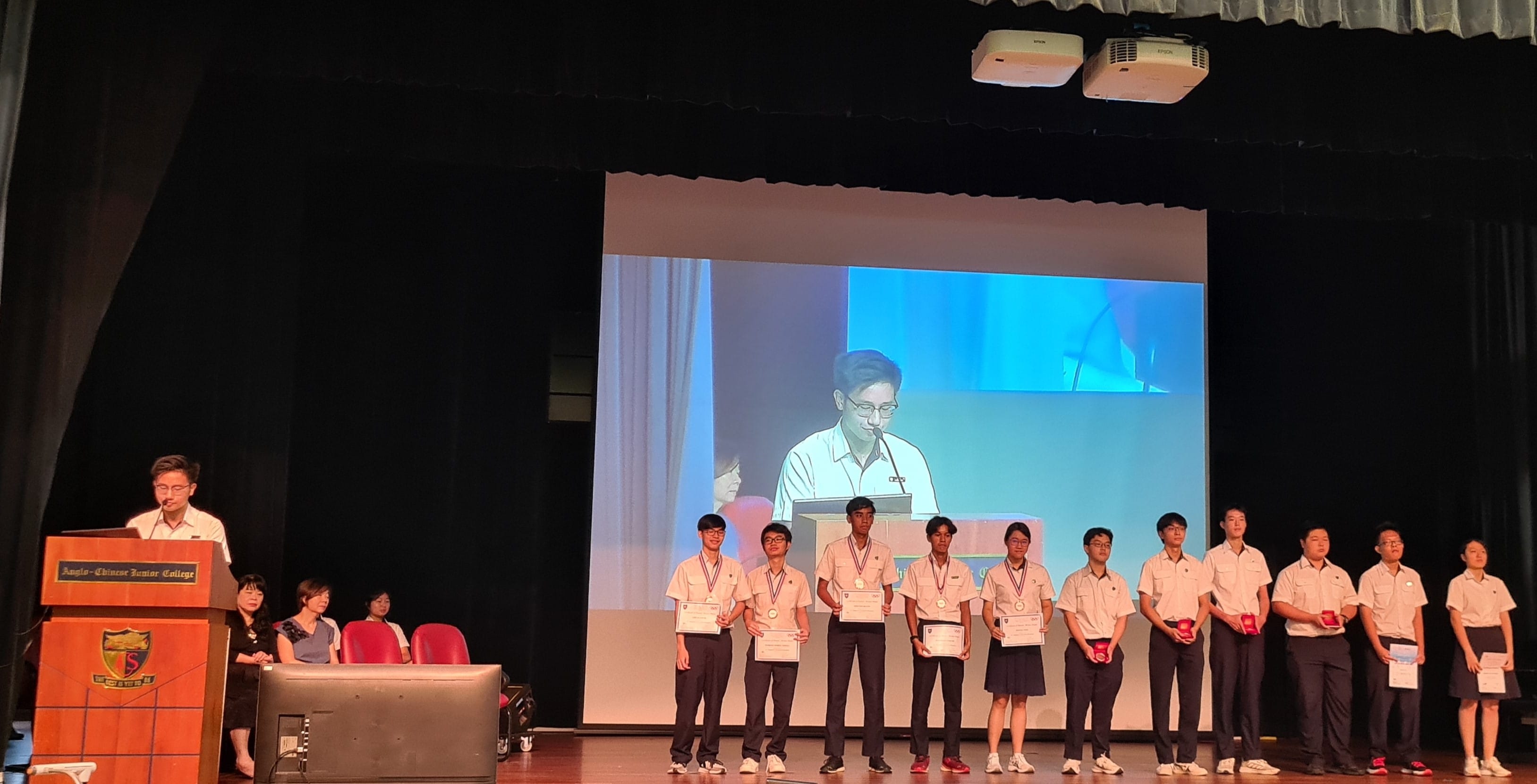 Students in uniforms stand on stage holding certificates and medals at an Anglo-Chinese Junior College awards ceremony.