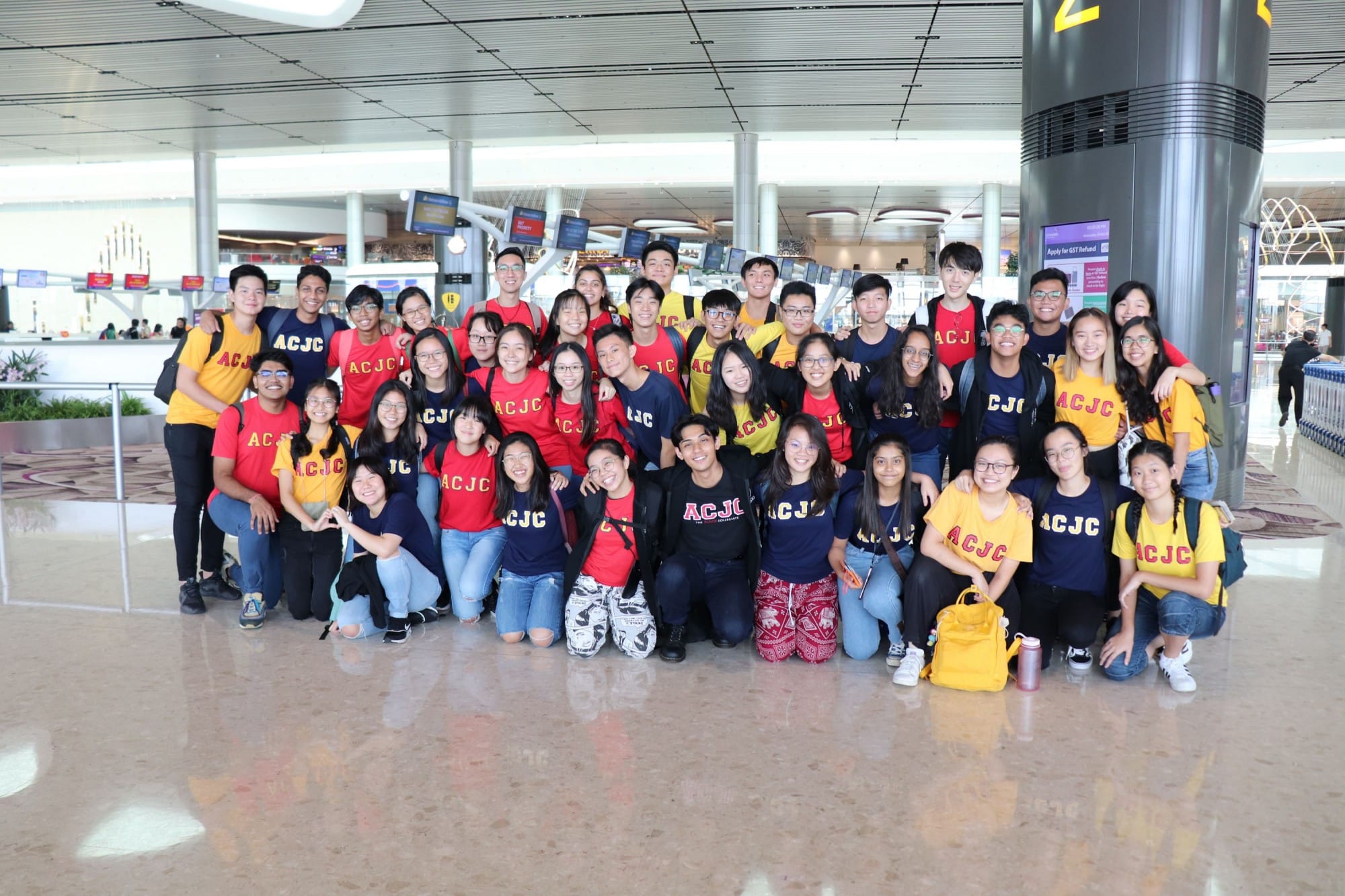 Large group of young people wearing red, navy, and yellow ACJC shirts posing together inside an airport terminal.