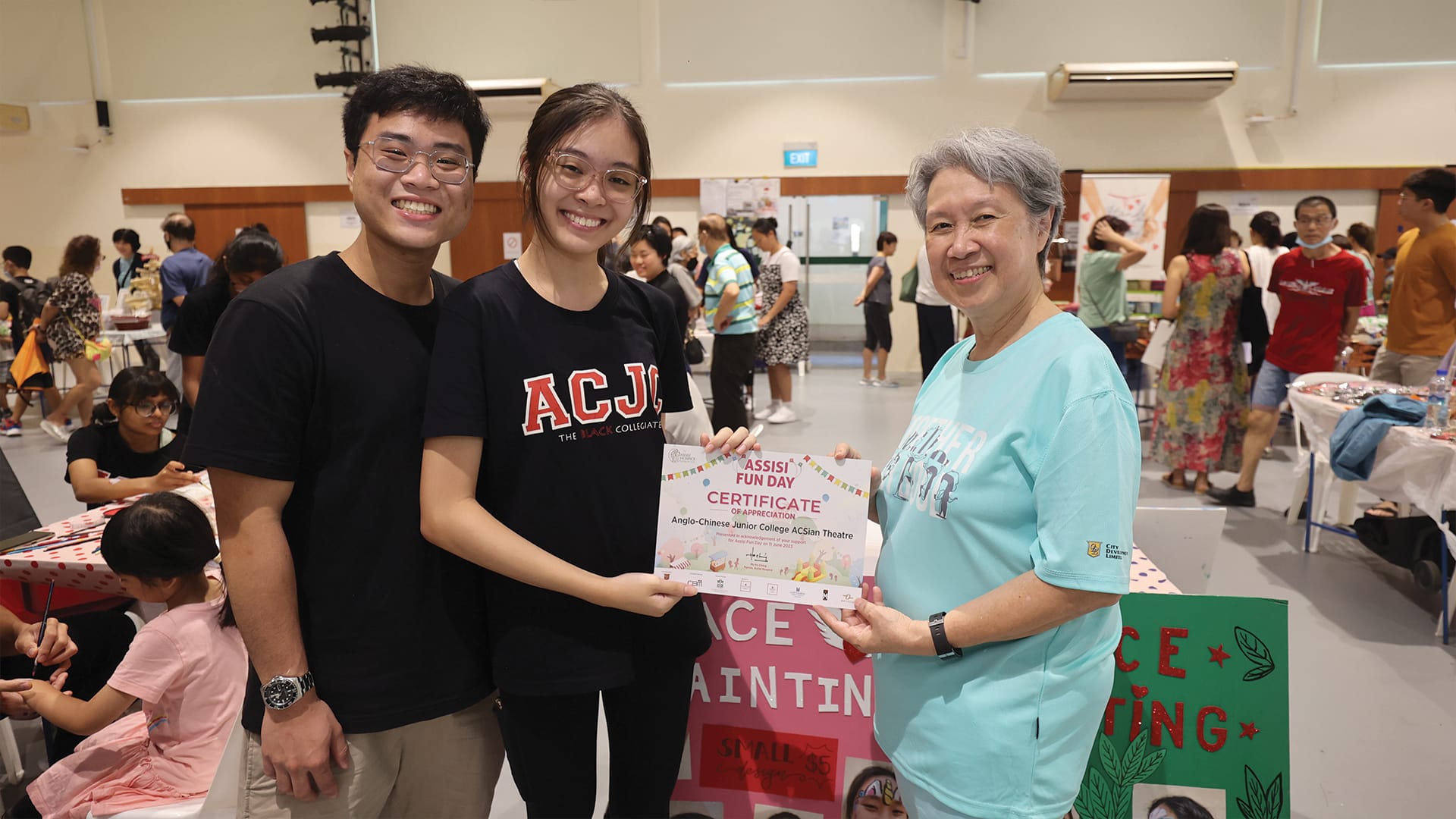 Three smiling people hold an Assisi Fun Day Certificate of Appreciation at a busy indoor community event.