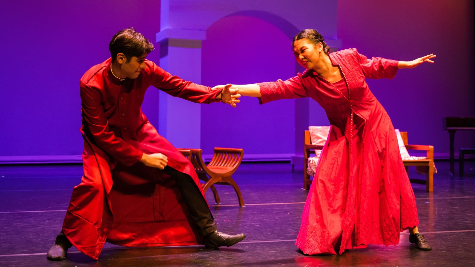 Two performers in red costumes hold hands dramatically on a purple-lit stage with wooden chairs in the background.
