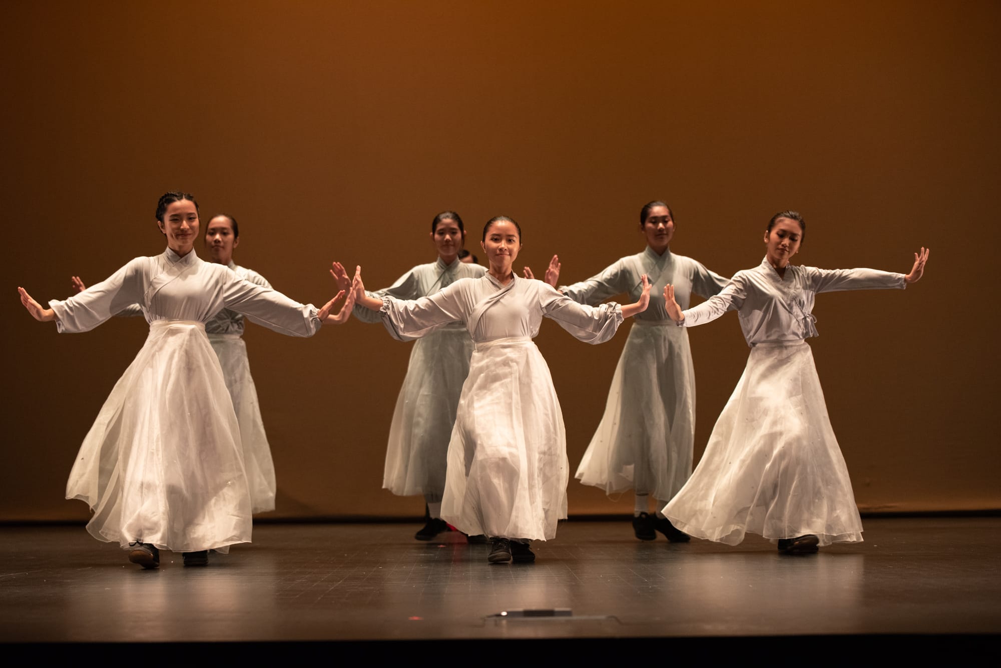 Seven women in white flowing skirts perform a synchronized traditional Chinese dance on a warmly lit stage.
