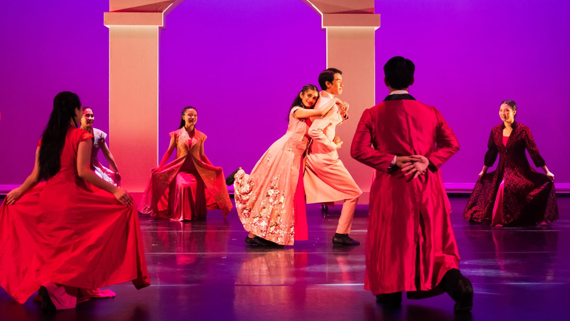 Performers in red and pink formal gowns and suits dance on a purple-lit stage with white arch backdrop.