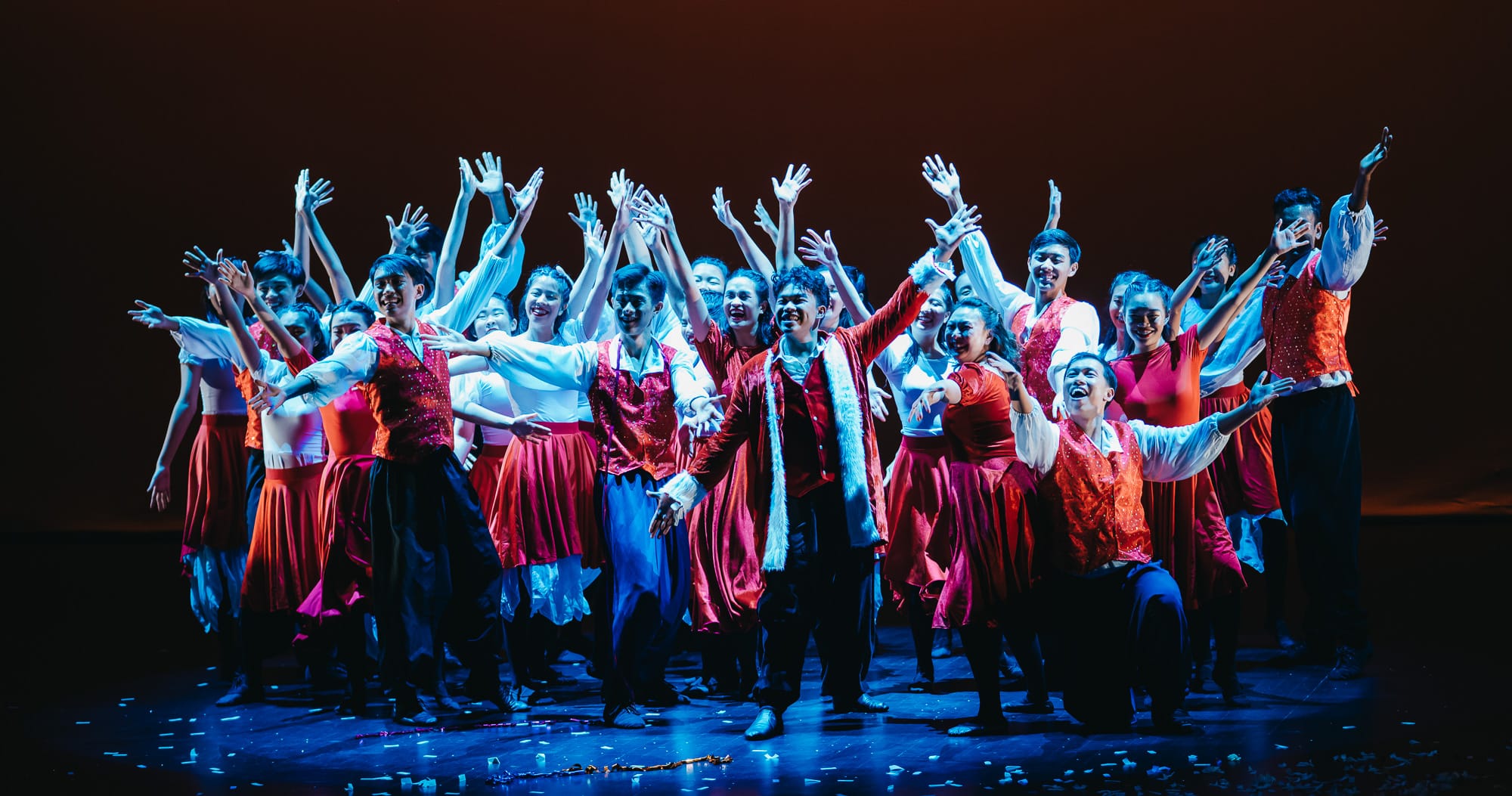 Large group of performers in red and white costumes dancing on a lit stage with arms raised.
