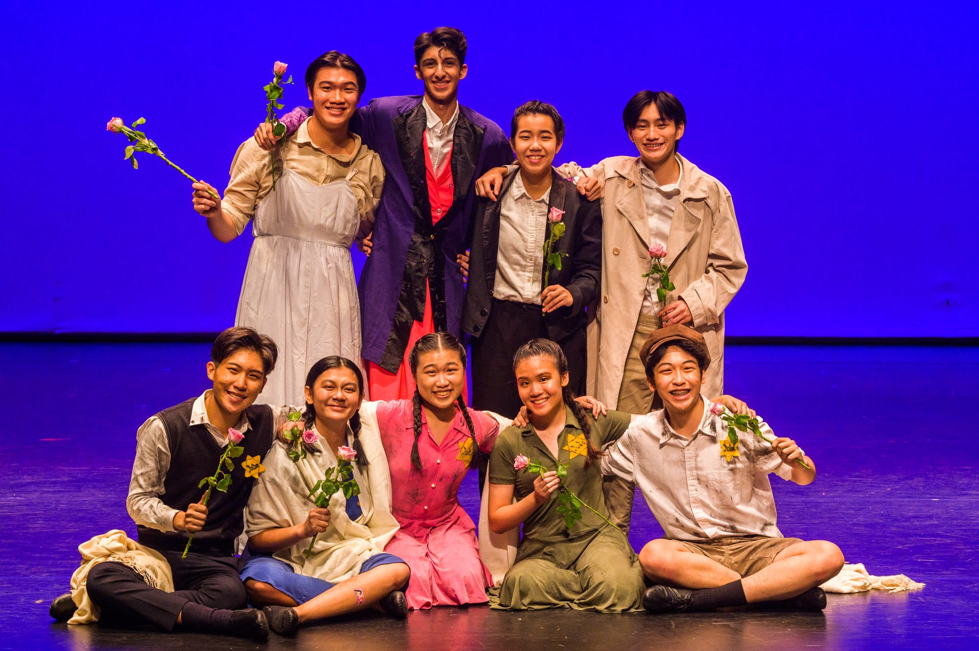 Nine young performers in period costumes pose on a blue-lit stage, holding pink roses and smiling.