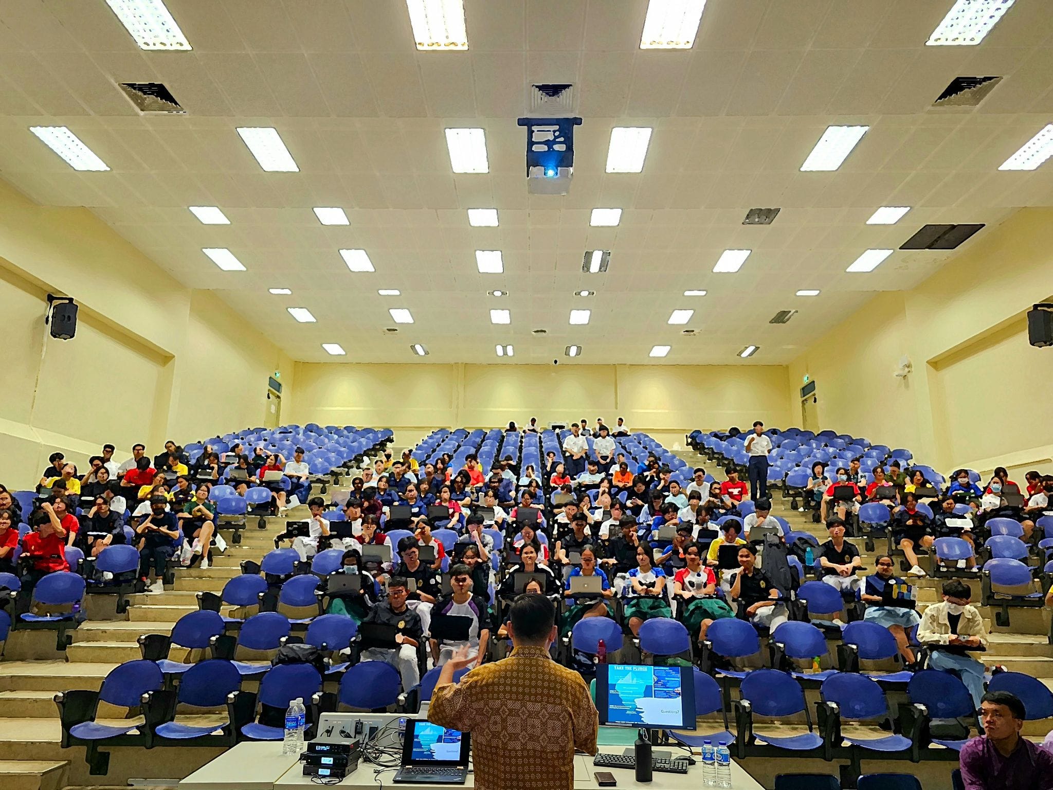 A speaker stands at the front of a large lecture hall filled with students seated in blue chairs.