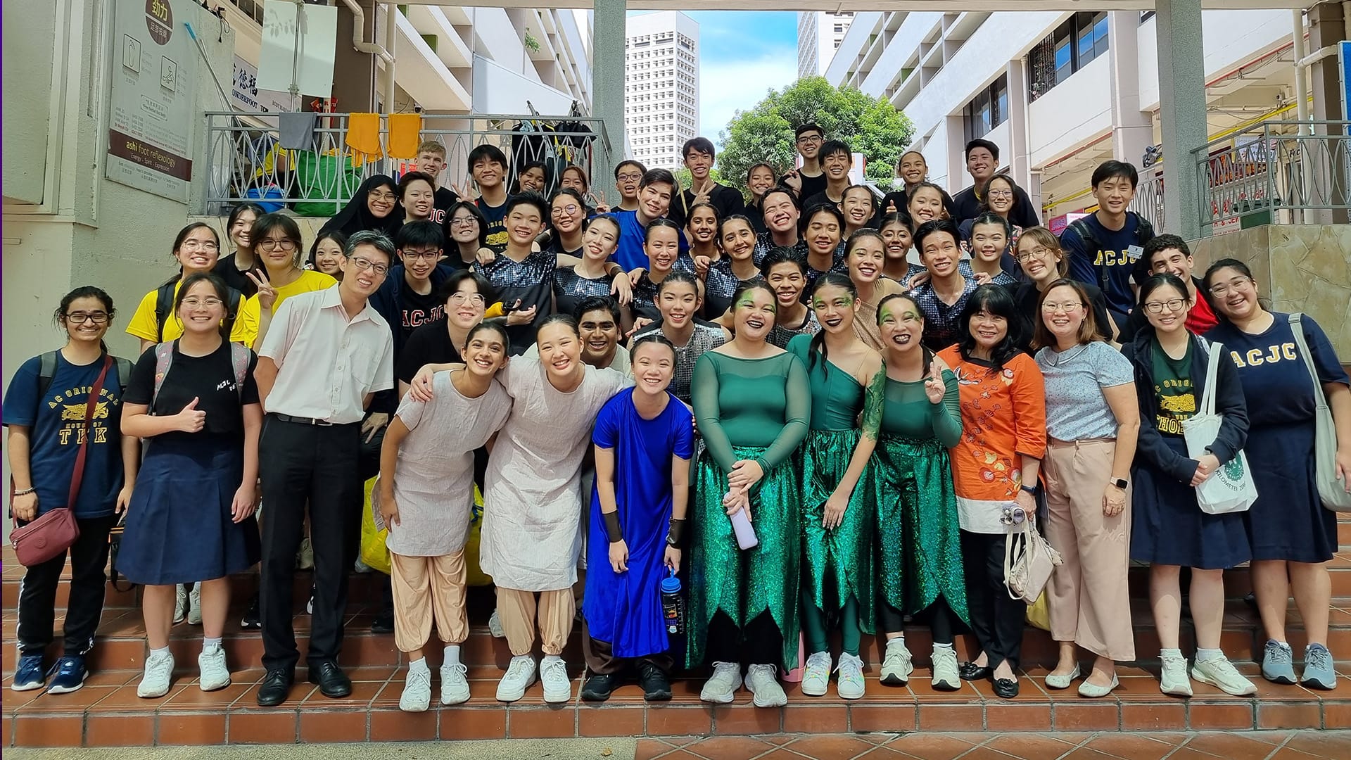 Large group of students in ACJC attire and colorful costumes posing together on outdoor steps.