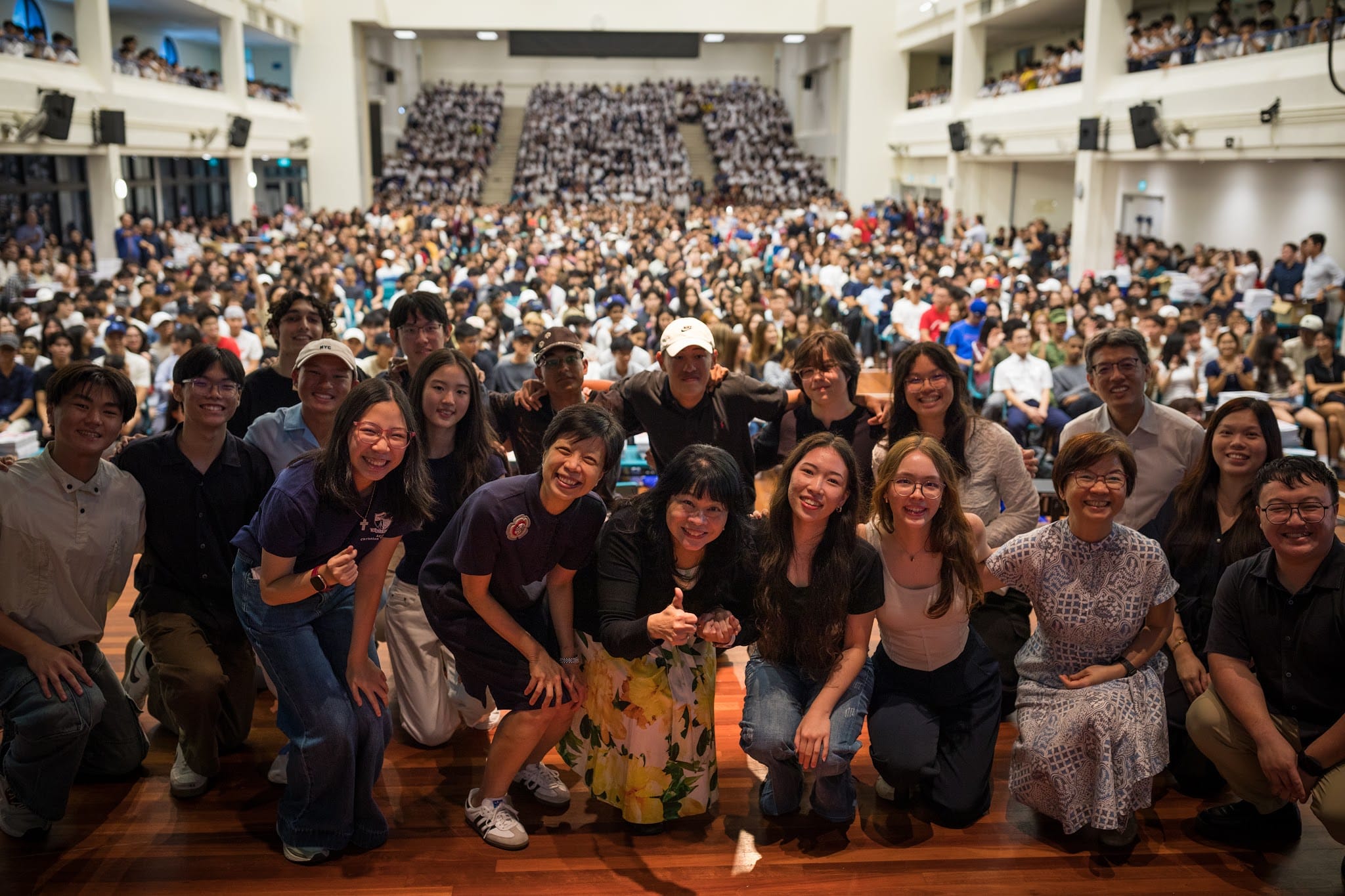 A large group poses for a photo on a stage in a packed auditorium filled with hundreds of seated attendees.