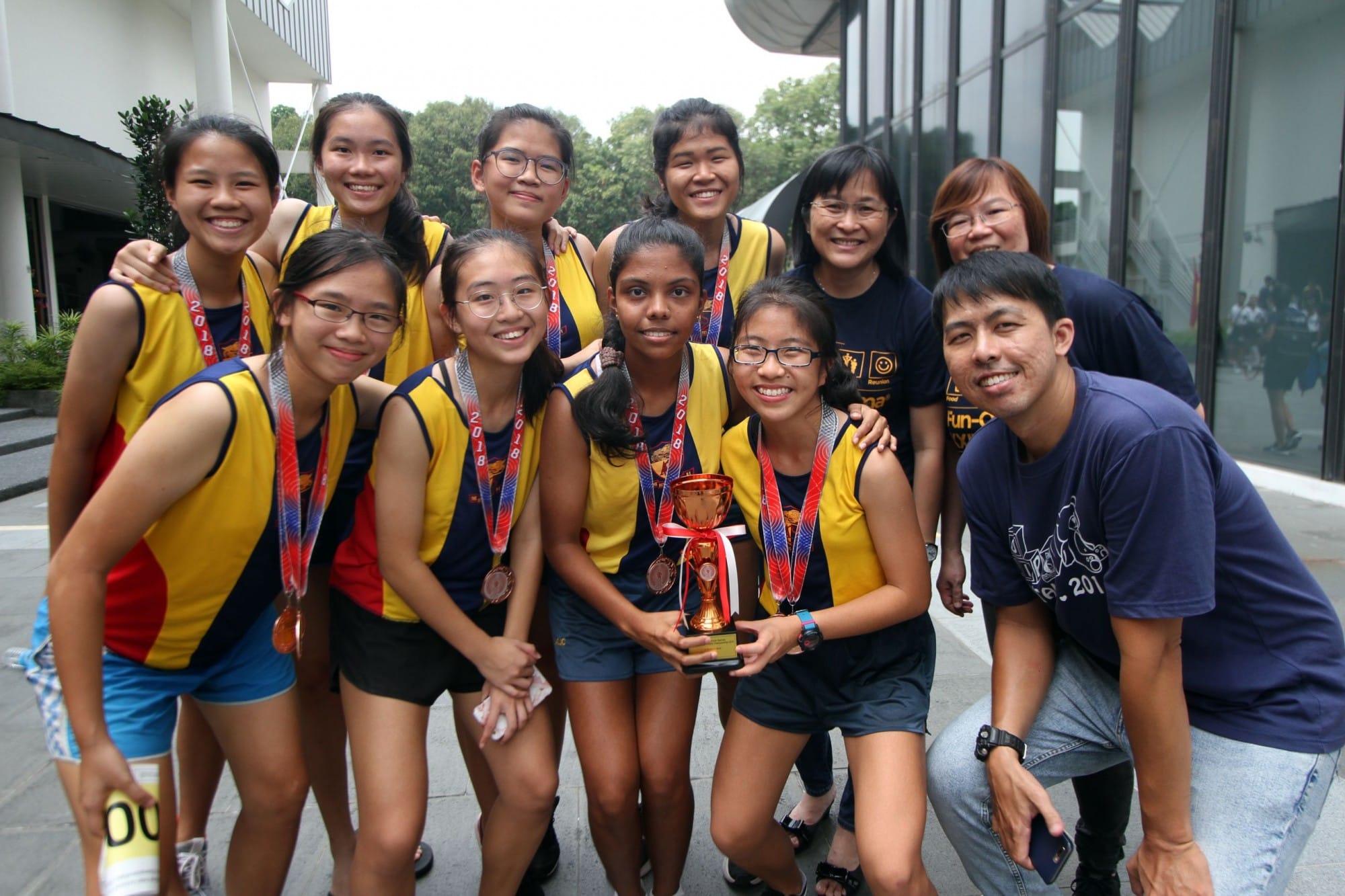 A group of smiling young girls in yellow and blue jerseys with medals pose with a trophy and three adults.