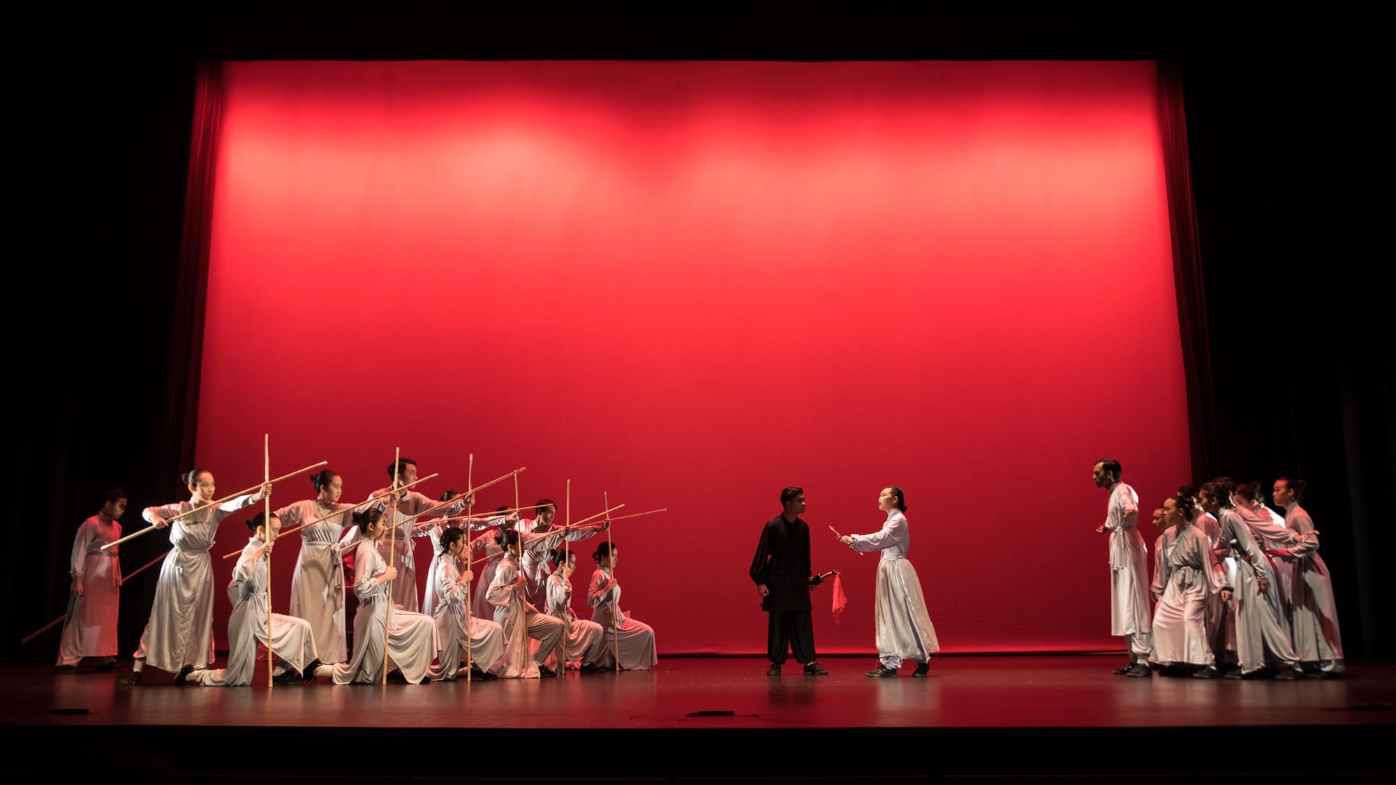 Performers in white robes hold wooden staffs on a stage lit by a bright red backdrop.