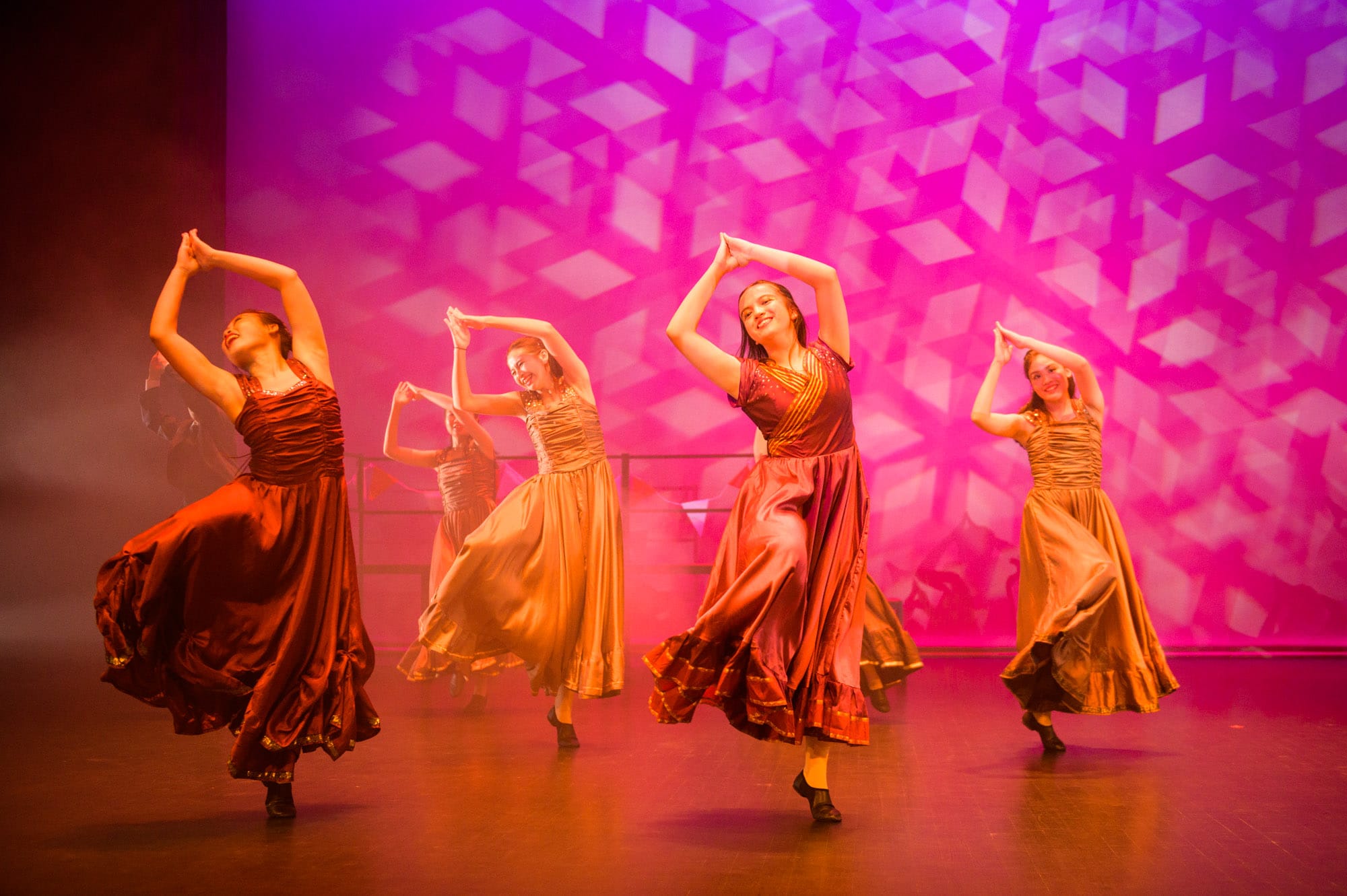 Four female dancers in flowing red and gold gowns perform on stage with arms raised before a pink geometric backdrop.