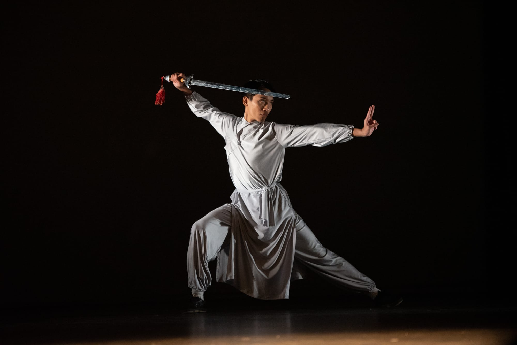 Man in white traditional outfit performs a martial arts stance, holding a sword overhead with a red tassel, against black background.