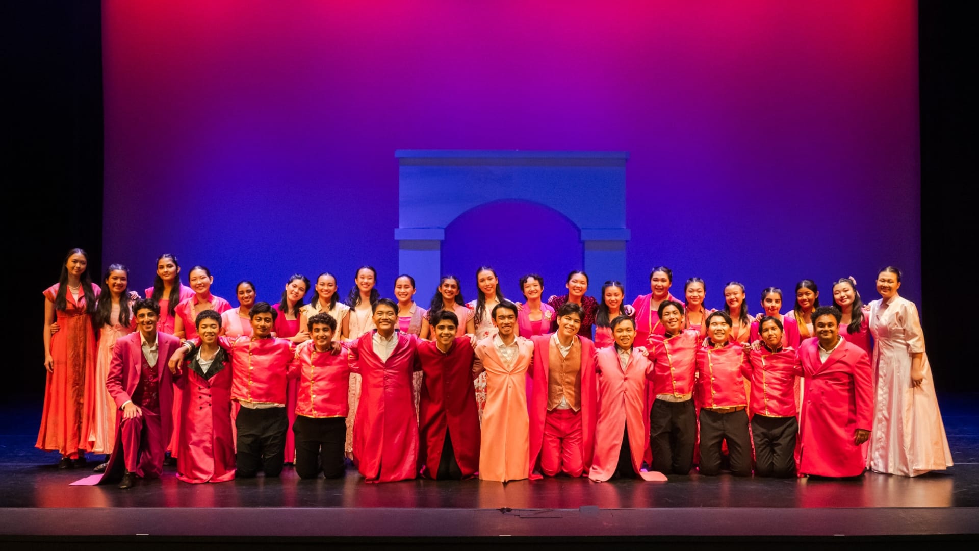 Large group of performers in pink and red costumes posing on a stage with a purple-lit backdrop and arch prop.
