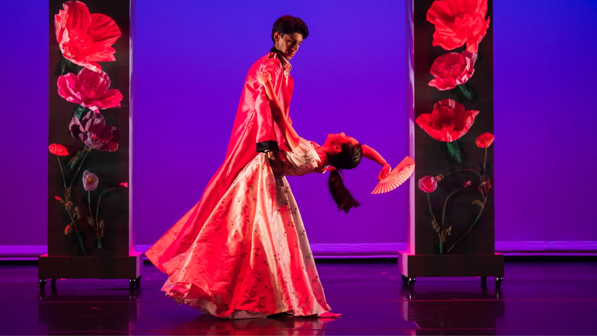 Two dancers in red traditional costumes perform a dip on a purple-lit stage with large red flower backdrops.
