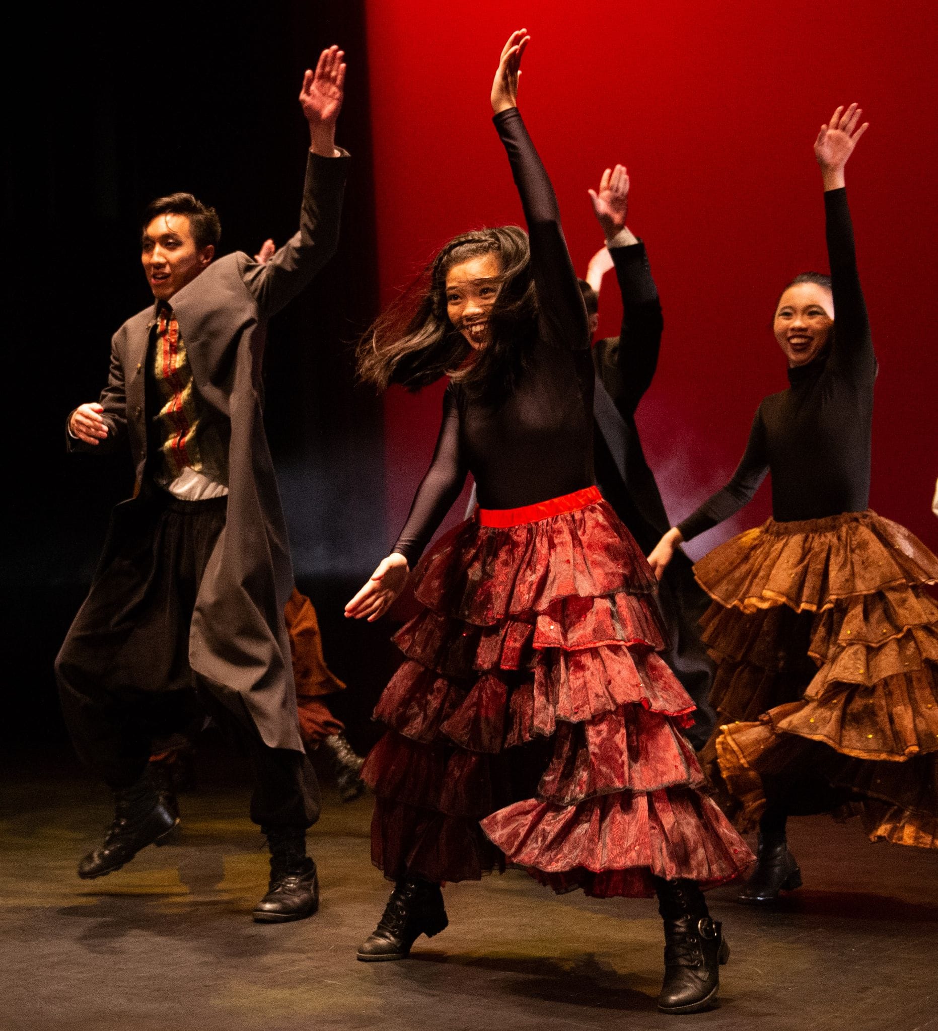 Dancers perform on stage, a girl in a red ruffled skirt raises her arms high against a red backdrop.