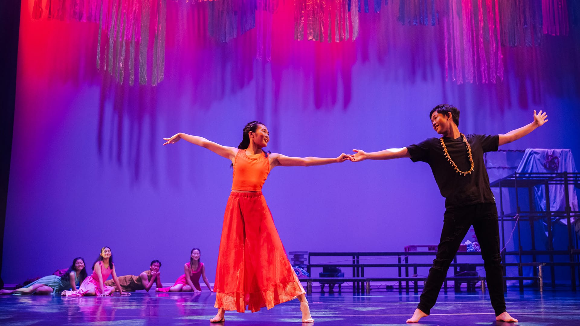 Two dancers hold hands on a lit stage, one in red dress, one in black, with colorful hanging streamers above.