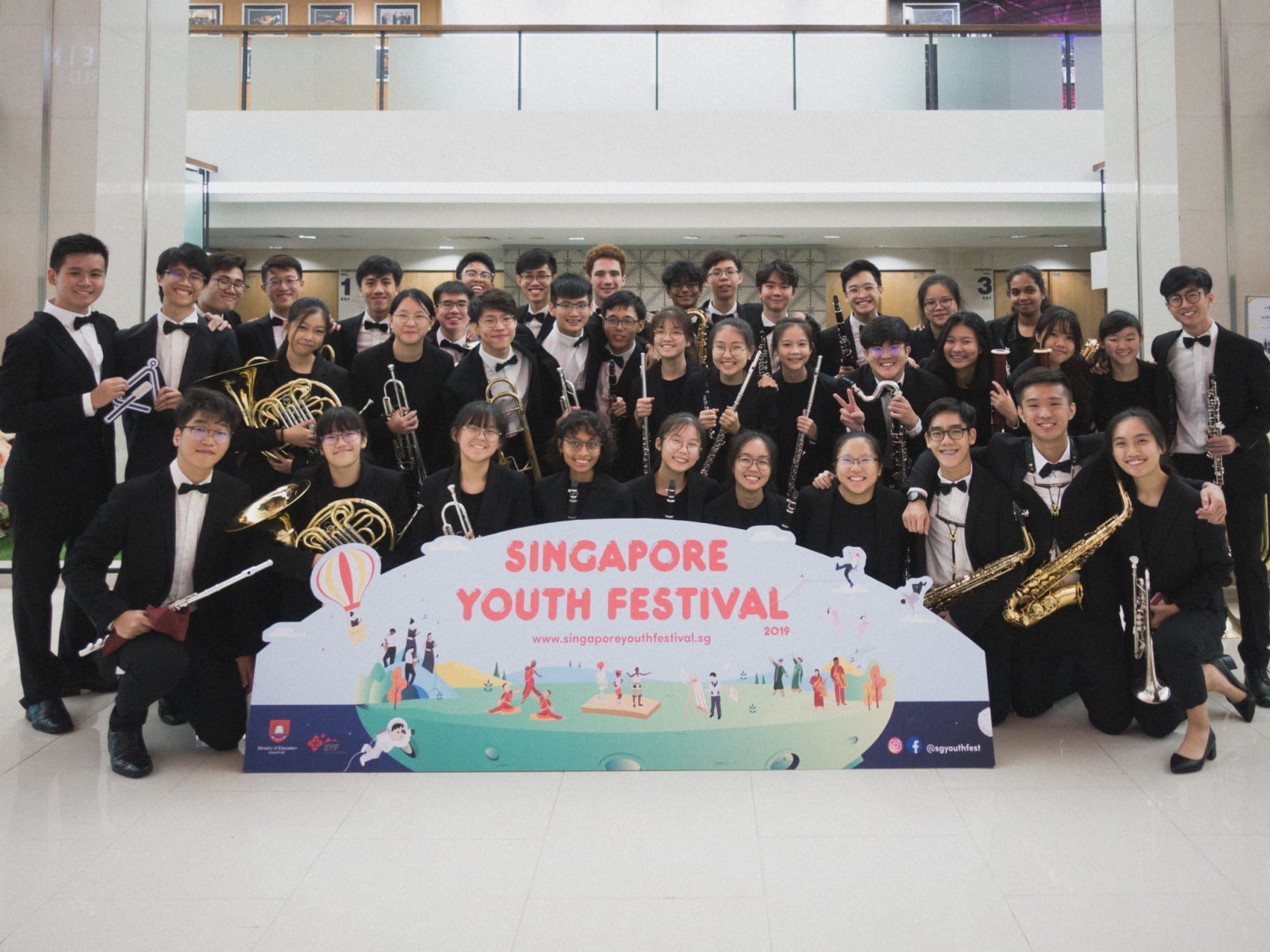 A youth orchestra in black formal wear poses with instruments behind a Singapore Youth Festival 2019 banner.