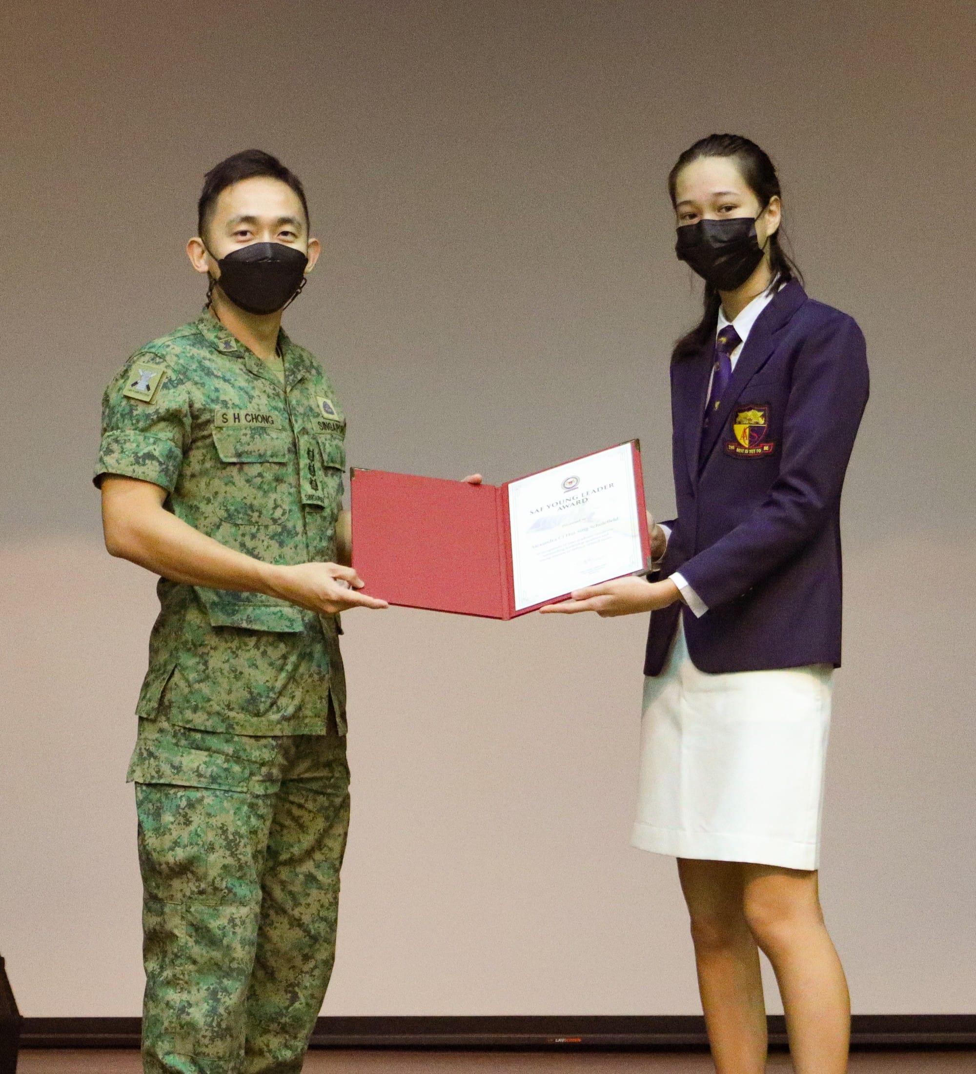 A soldier in camouflage uniform and a student in a purple blazer both wearing masks hold a certificate together.