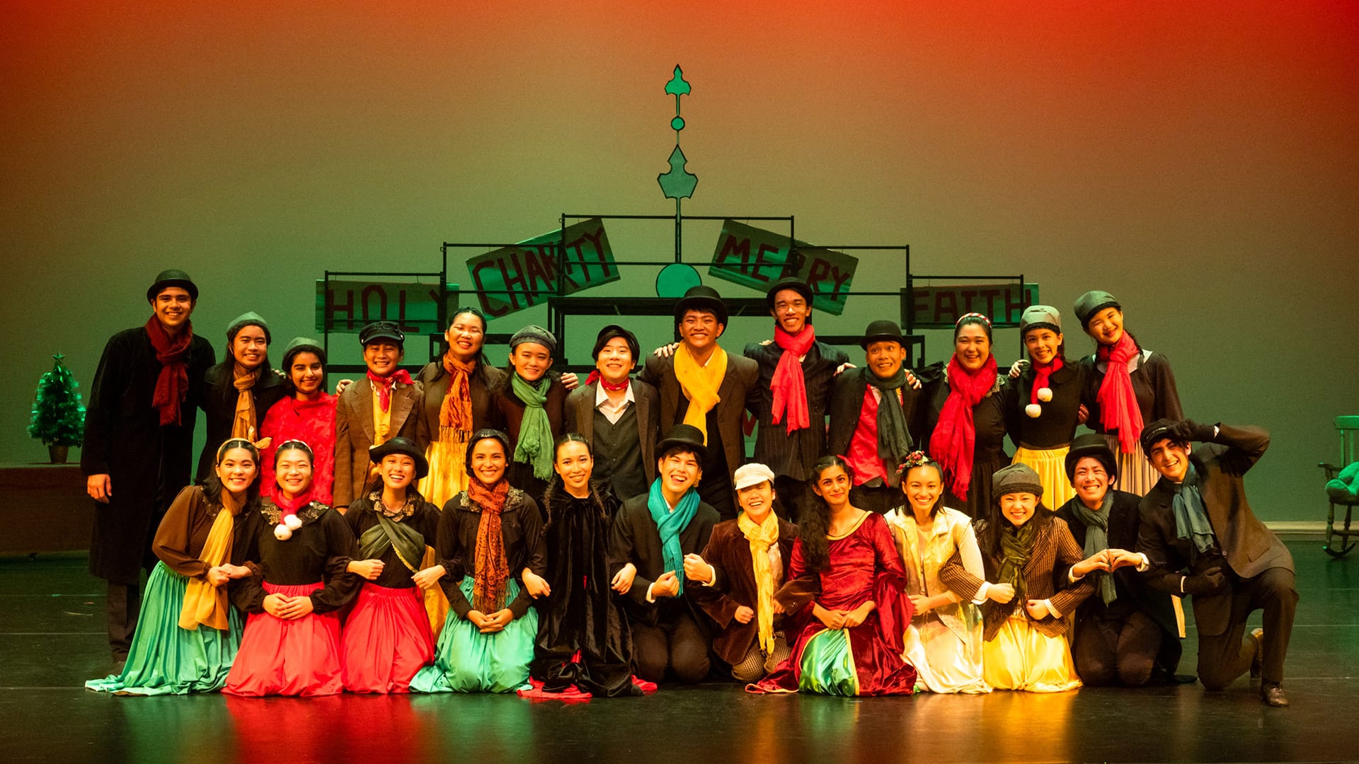 A large cast in colorful Victorian costumes poses on a stage with a holiday-themed backdrop reading "Holy Charity Merry."
