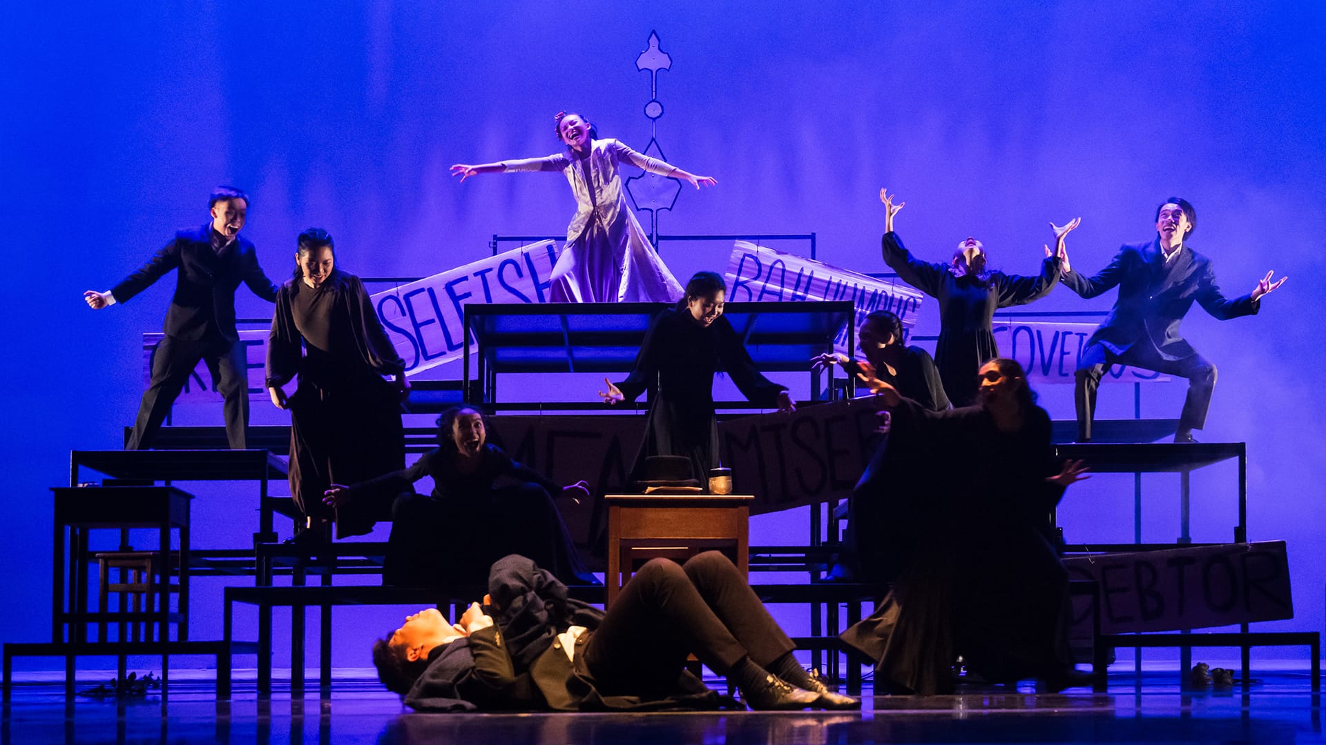 Stage performers in black costumes pose dramatically on tiered platforms under blue lighting, with one in white standing elevated.