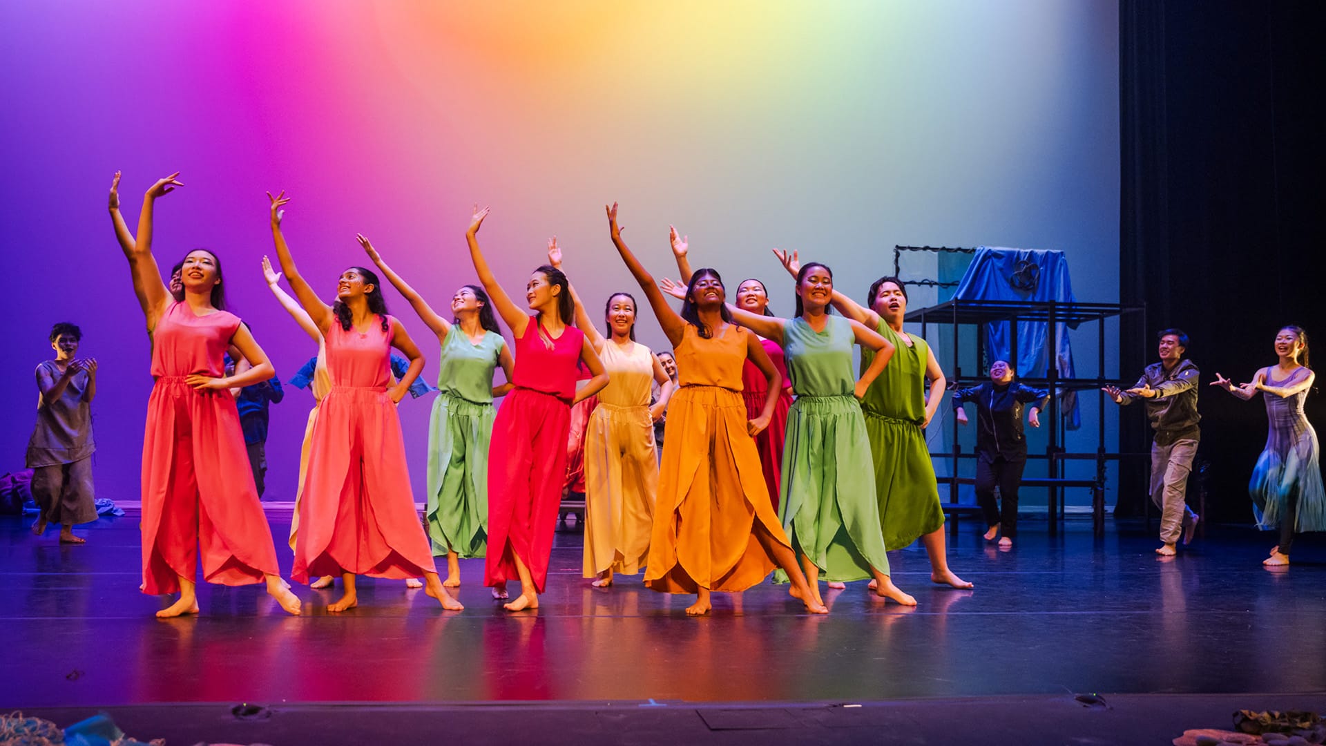 Dancers in colorful flowing outfits perform on a lit stage with rainbow-colored backdrop, arms raised.