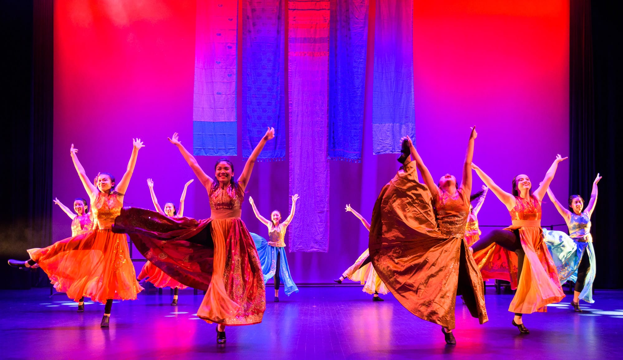 Dancers in colorful flowing skirts perform on a stage lit in red and purple, arms raised, with blue fabric backdrops.