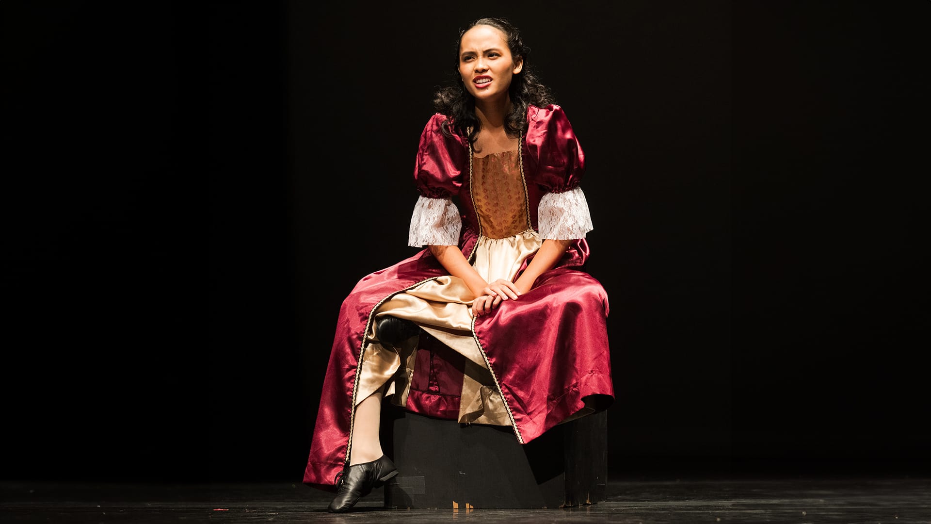 A woman in a red and gold period costume sits on a dark stage, mouth open as if speaking or singing.