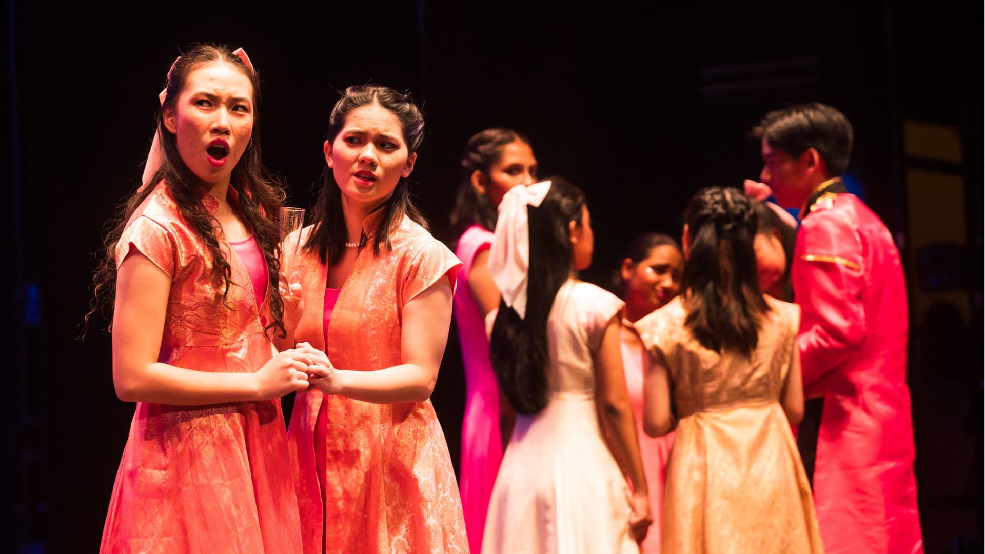 Two women in matching pink brocade dresses sing on stage while other performers in colorful costumes stand behind them.