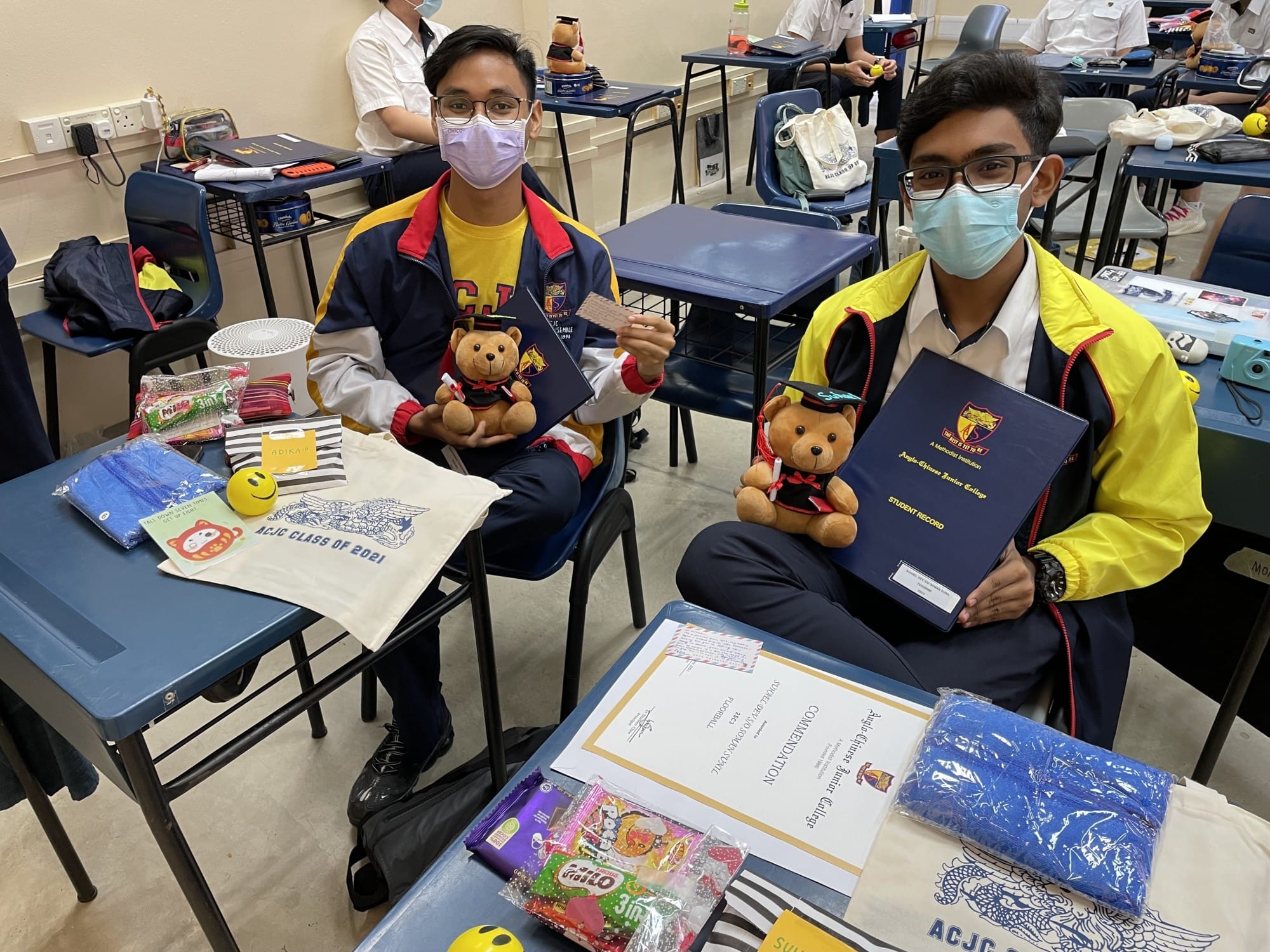 Two masked students in school uniforms hold graduation teddy bears and folders, seated at desks with gifts and a commendation certificate.