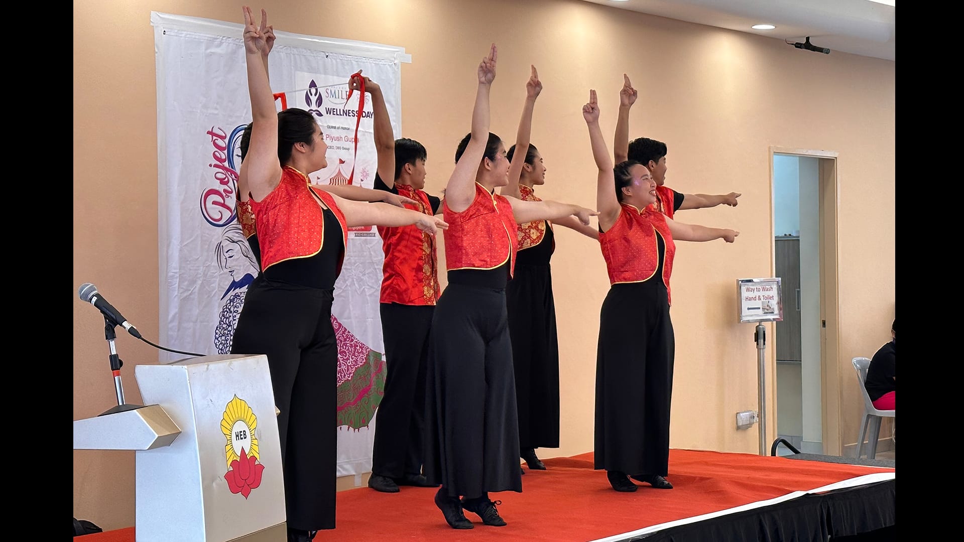 Six performers in red and black outfits dance on a red stage with arms raised at an indoor wellness event.