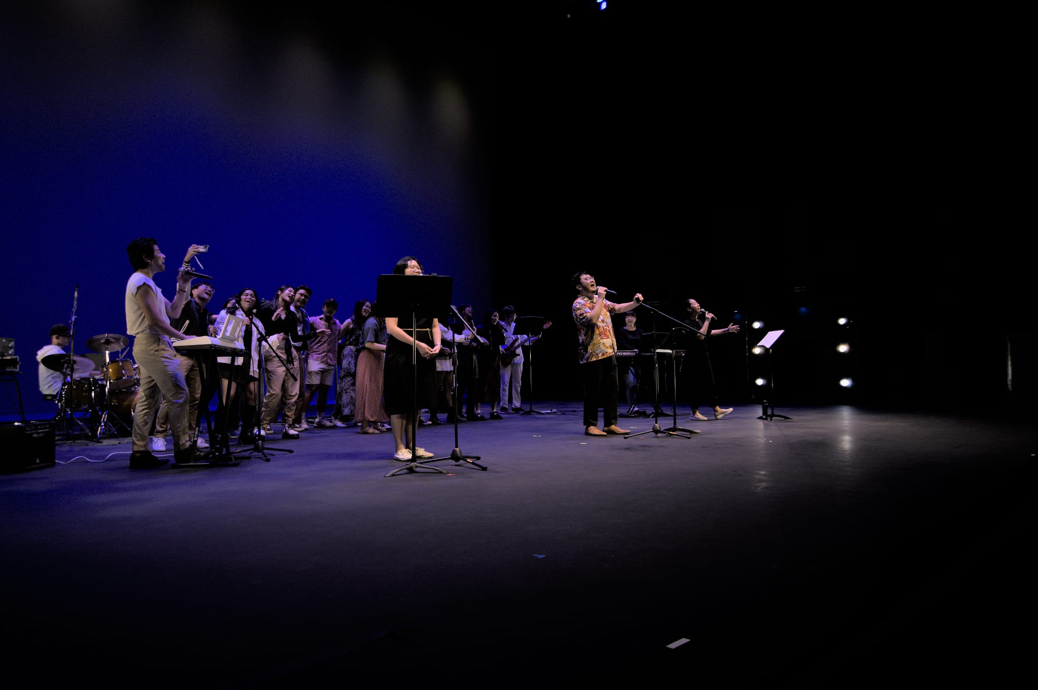 A group of performers on a dimly lit blue stage singing and playing instruments with music stands.