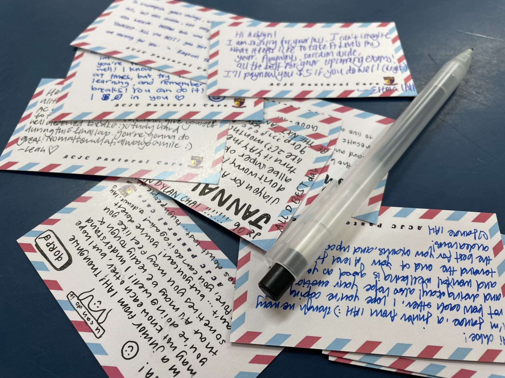 Several handwritten postcards with red and blue borders from ACJC Pastoral Care, with a pen resting on top.