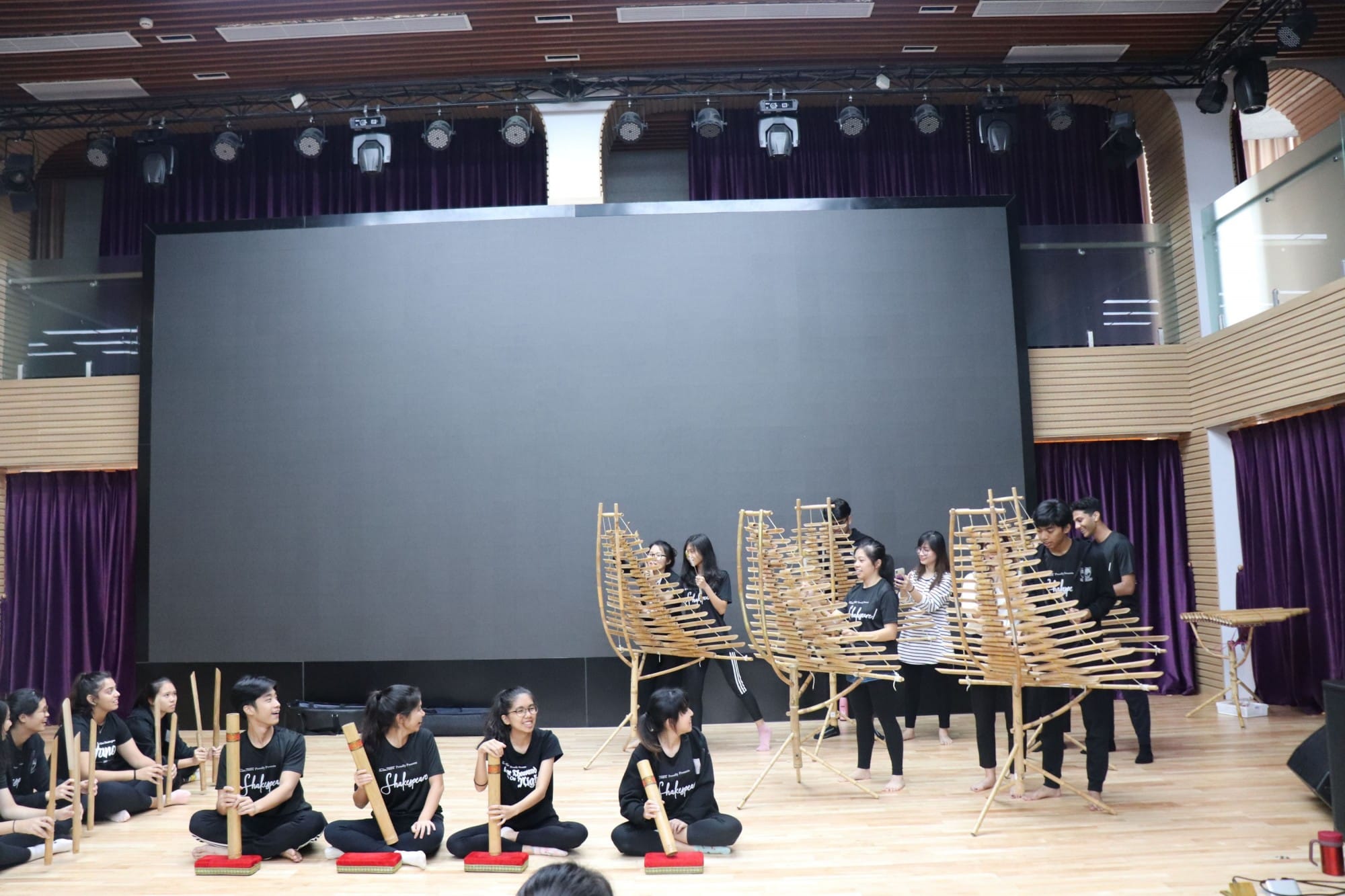 Group of young people in black shirts playing traditional bamboo instruments on a wooden stage floor.