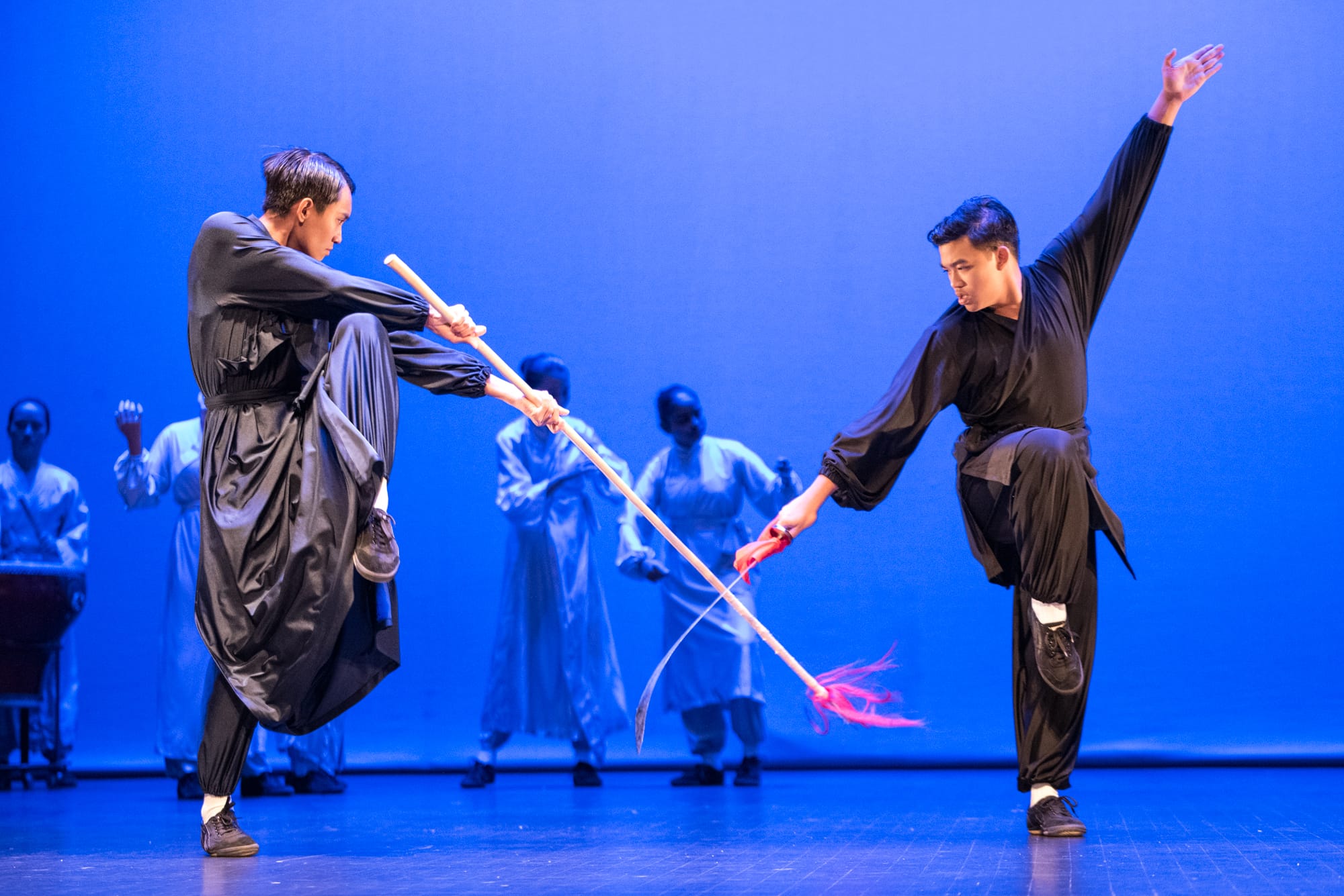 Two performers in black martial arts attire spar with a wooden staff on a blue-lit stage with others in background.